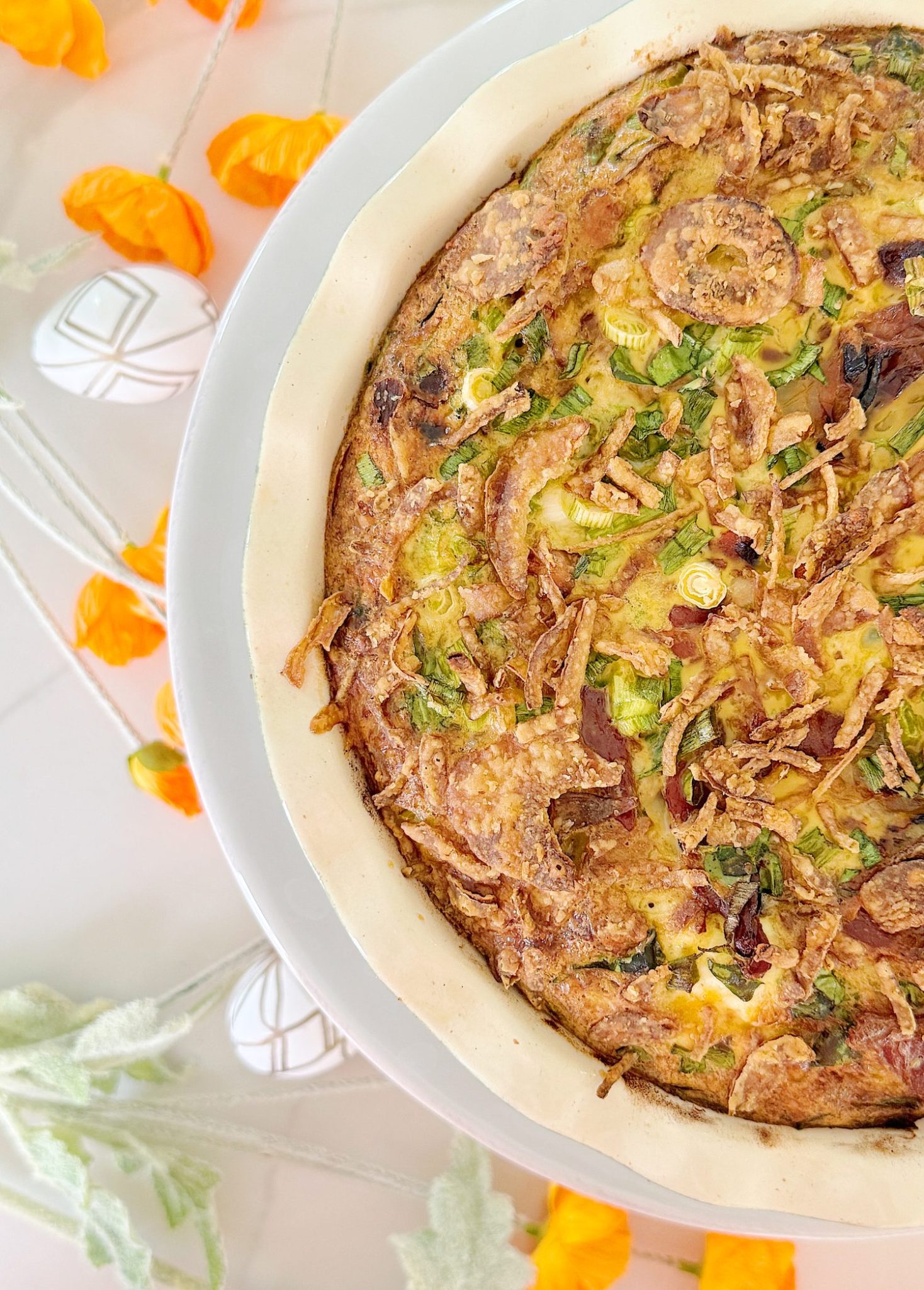 A savory pie with a crispy crust, topped with green onions, crispy fried onions, and sliced vegetables on a white plate. Orange flowers and decorative items surround the dish.