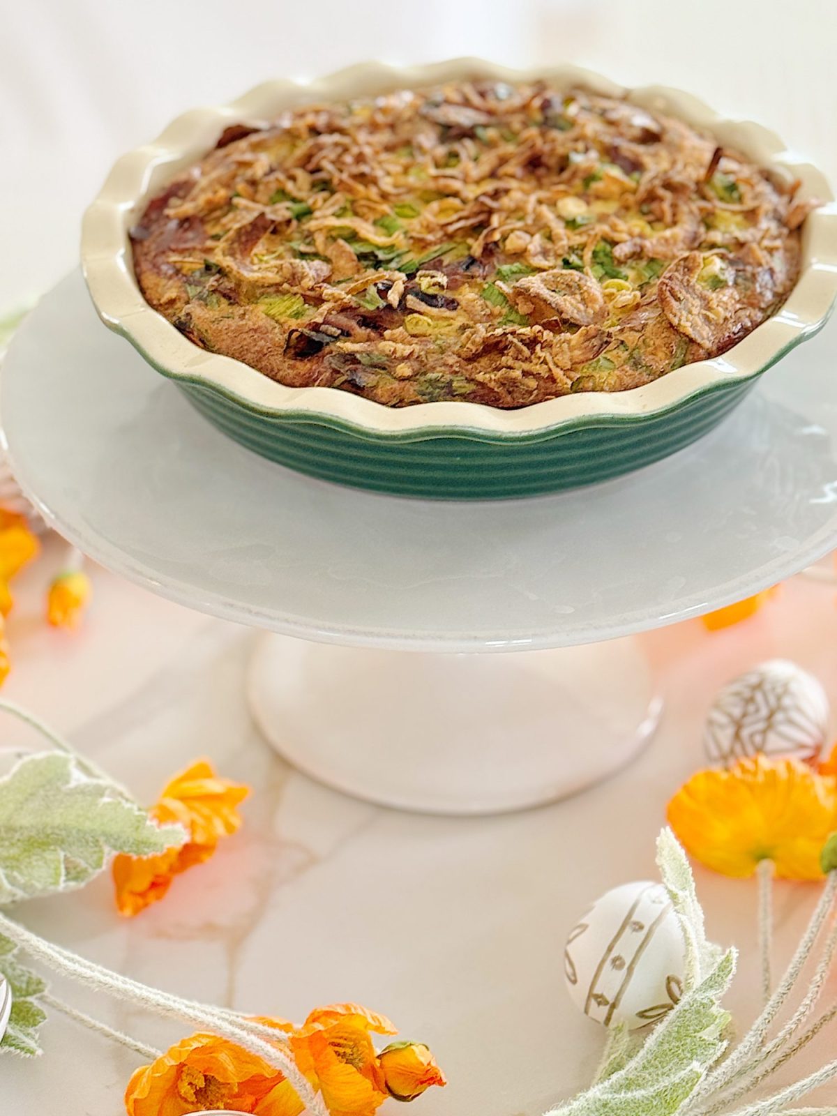 A savory quiche with a golden crust sits on a white stand, surrounded by orange flowers and decorative elements on a marble surface.