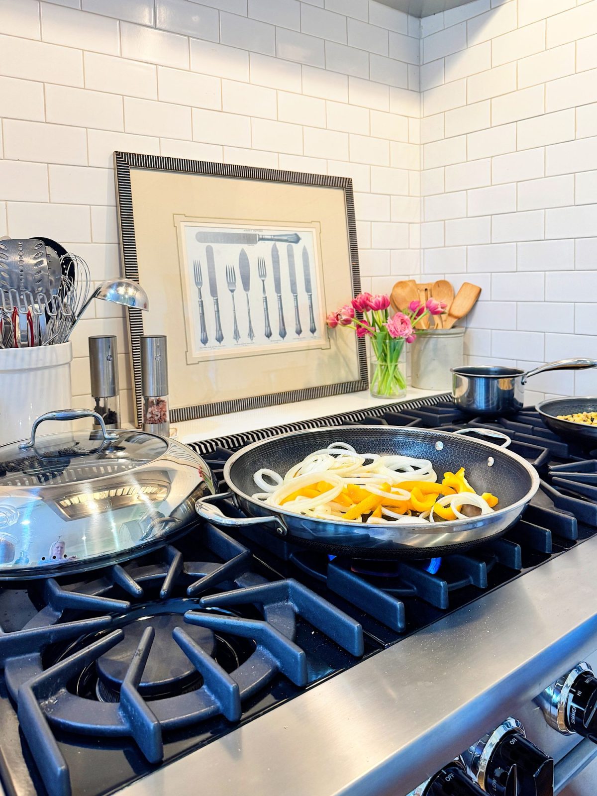 A stove with pans cooking sliced onions and bell peppers. Utensils, pink flowers, and framed cutlery art are on a countertop with white tile backsplash.