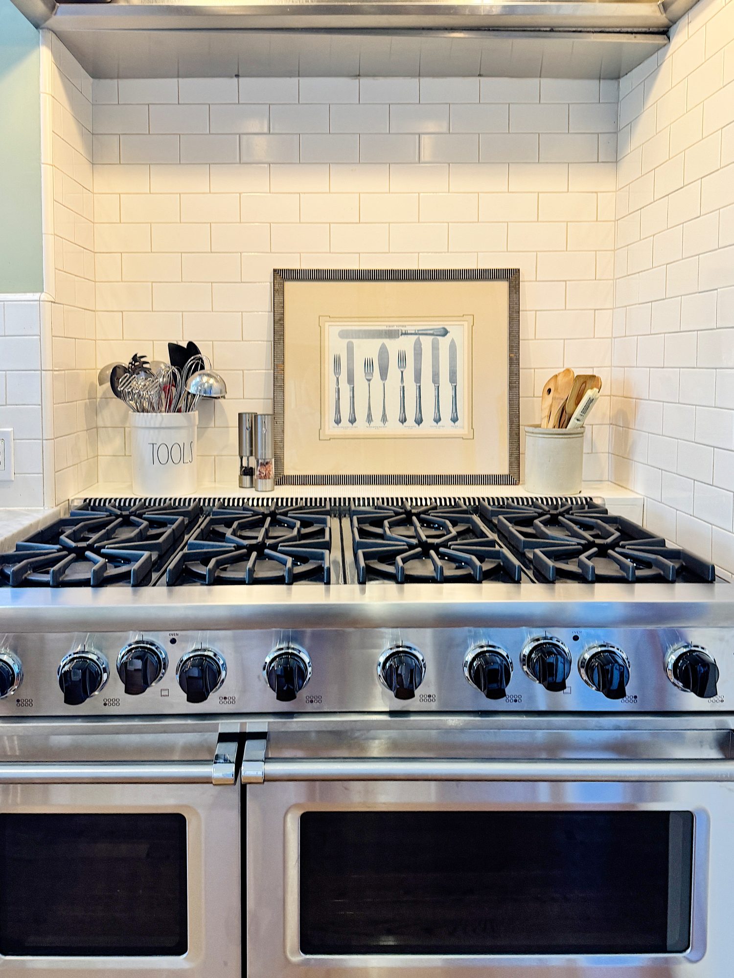 A stainless steel stove with six burners and two ovens. Above it is a framed utensil artwork against a white subway tile backsplash, flanked by kitchen tools and a utensil holder.