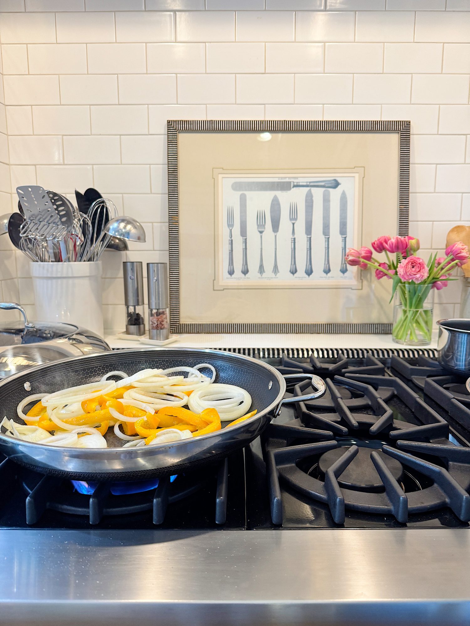 Sliced onions and bell peppers cooking in a pan on a stove. A framed utensil illustration, utensil holder, pink flowers, and a pepper mill are in the background.