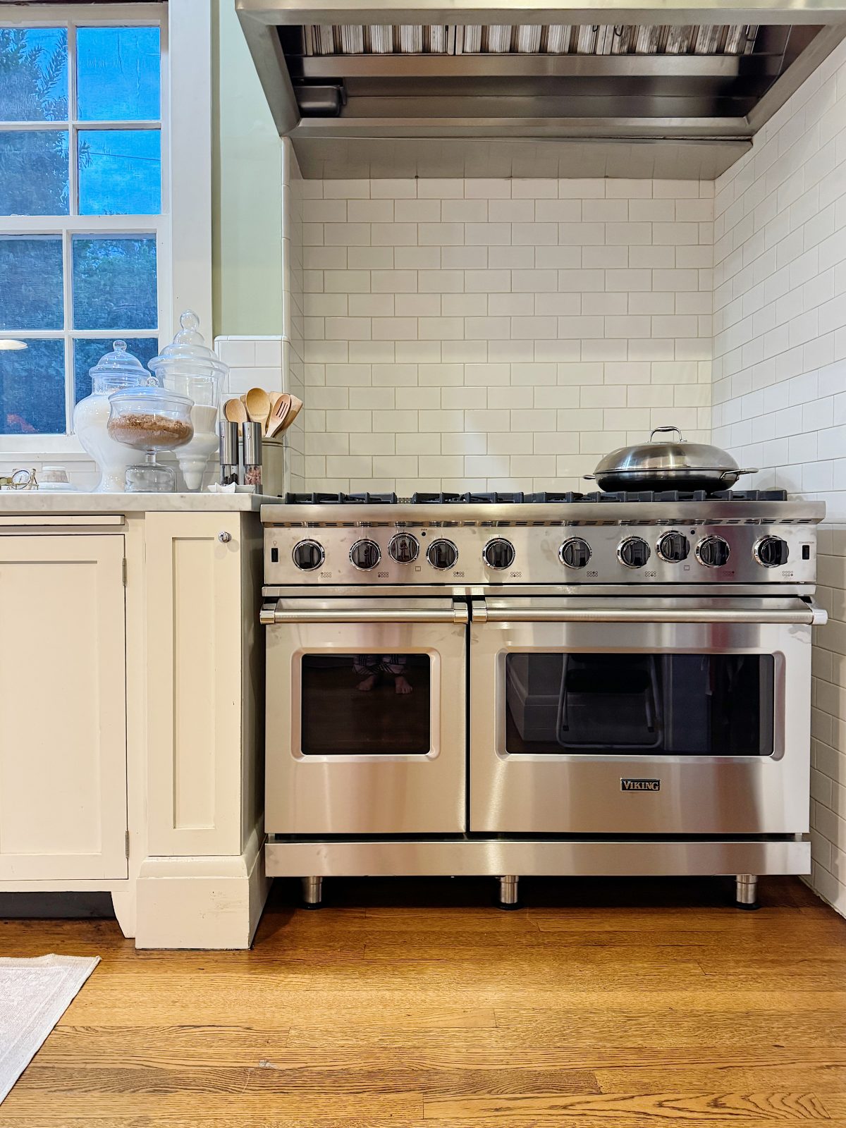 Stainless steel oven with vent hood and stove, next to white cabinetry and a window. Cookie jar and cooking utensils are on the counter. Wooden floor beneath.