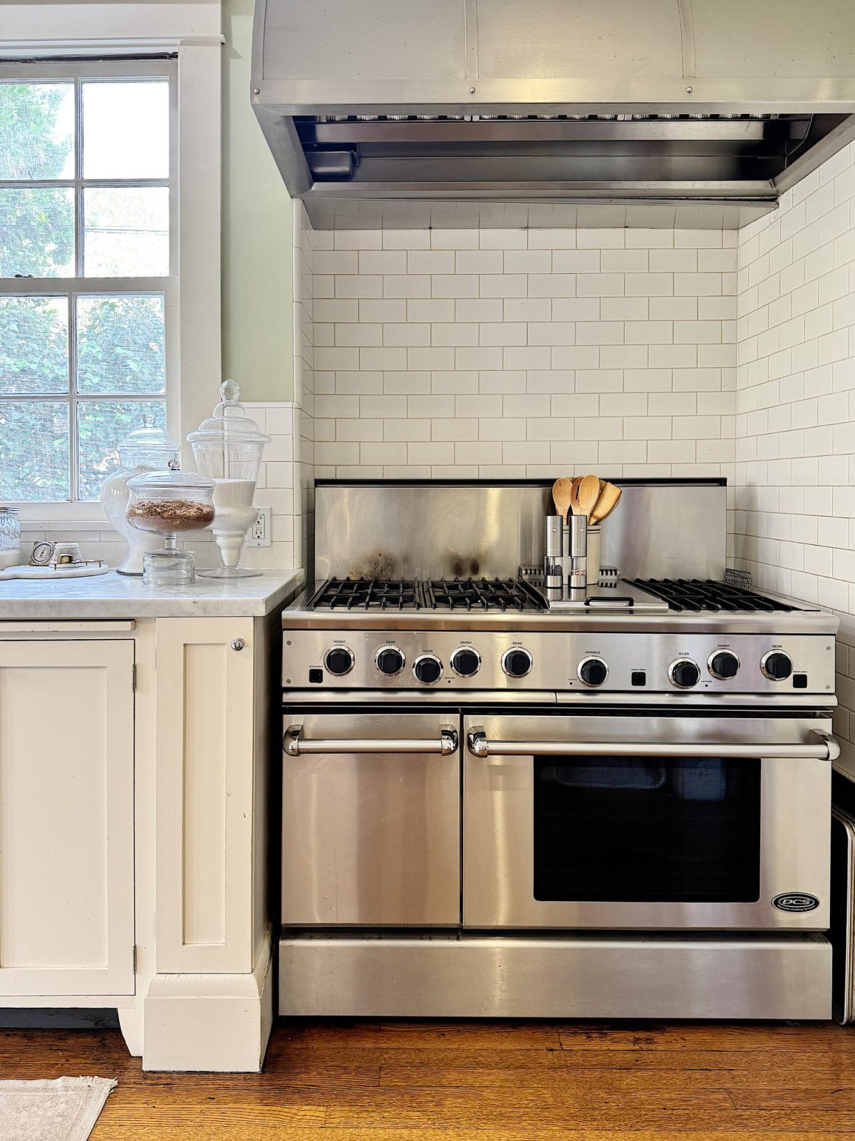 Modern kitchen with stainless steel stove and range hood. White subway tile backsplash and adjacent window. Countertop with cookie jars and utensils in a holder next to the stove.