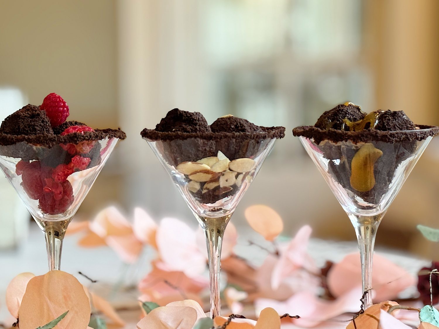 Three martini glasses filled with layered desserts, including raspberries and chocolate crumbles, almonds, and caramel sauce, surrounded by pink paper decorations.
