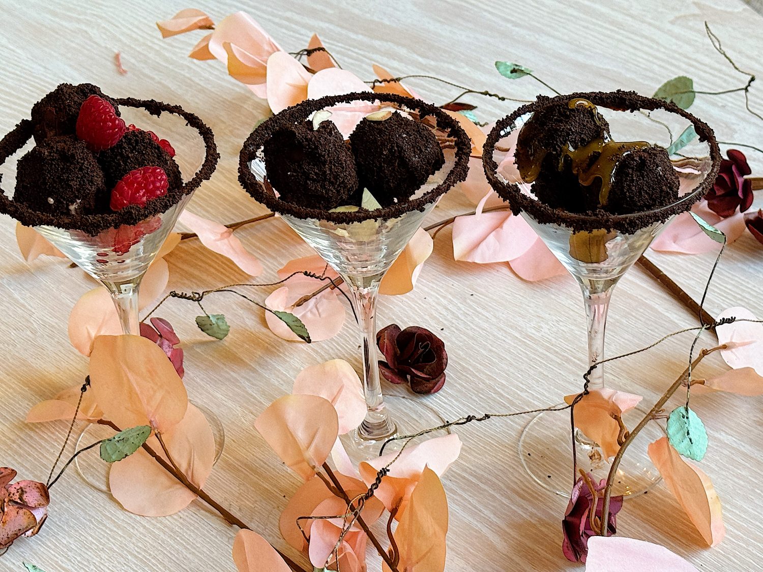 Three martini glasses with chocolate truffles, one garnished with raspberries, one with caramel drizzle, surrounded by decorative pink and brown leaves on a light wood surface.