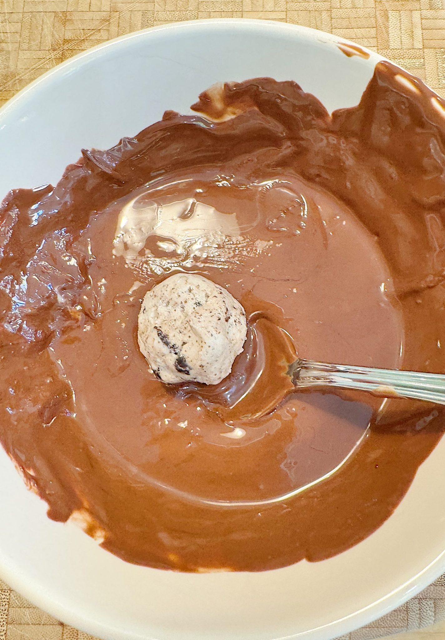 A ball of cookie dough rests in a bowl of melted chocolate with a spoon.
