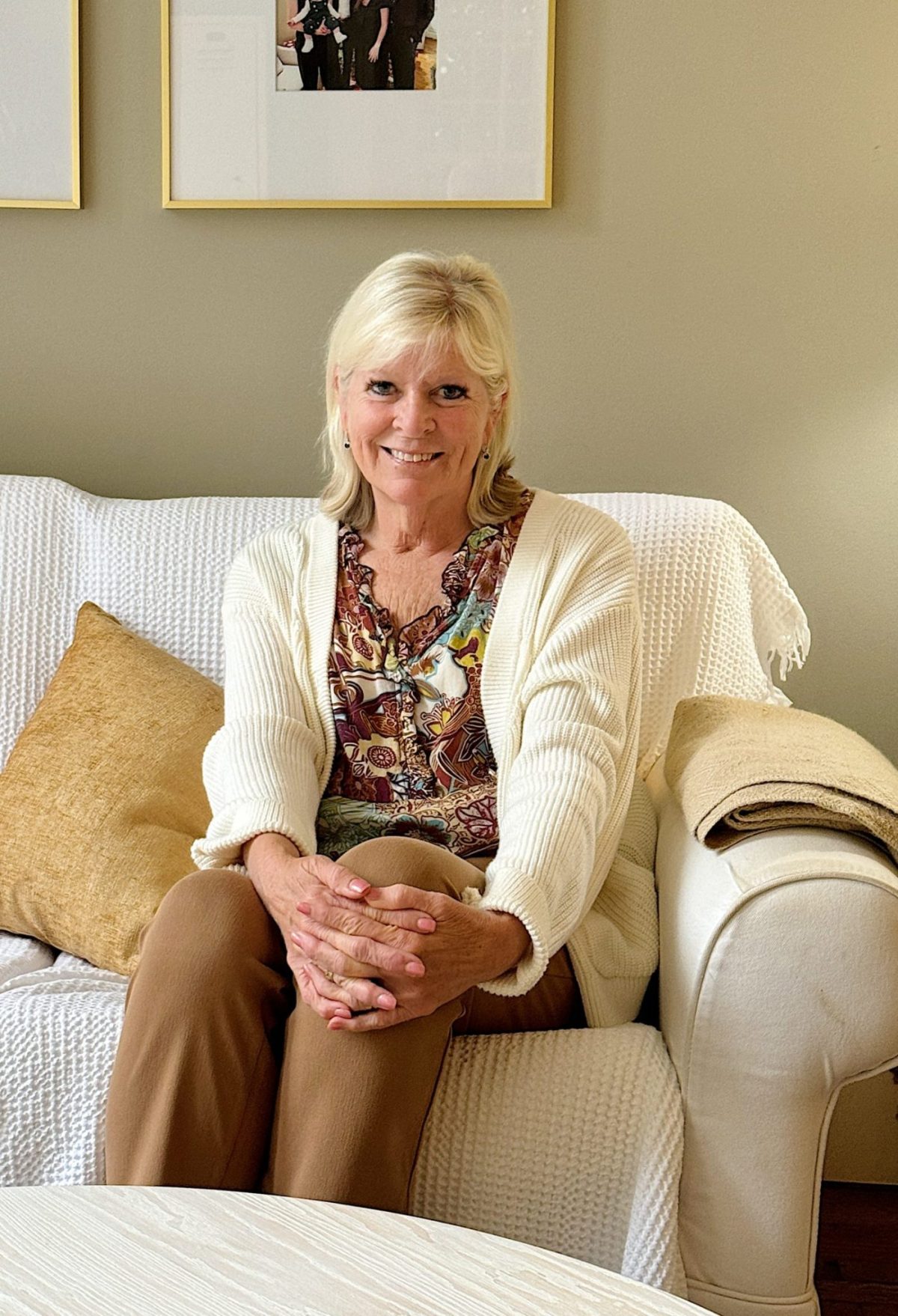 A woman sits on a white sofa, wearing a patterned blouse and cream cardigan, paired with light-colored trousers. She smiles warmly, reminiscent of a relaxed Amazon Prime evening, with framed photos decorating the wall behind her.