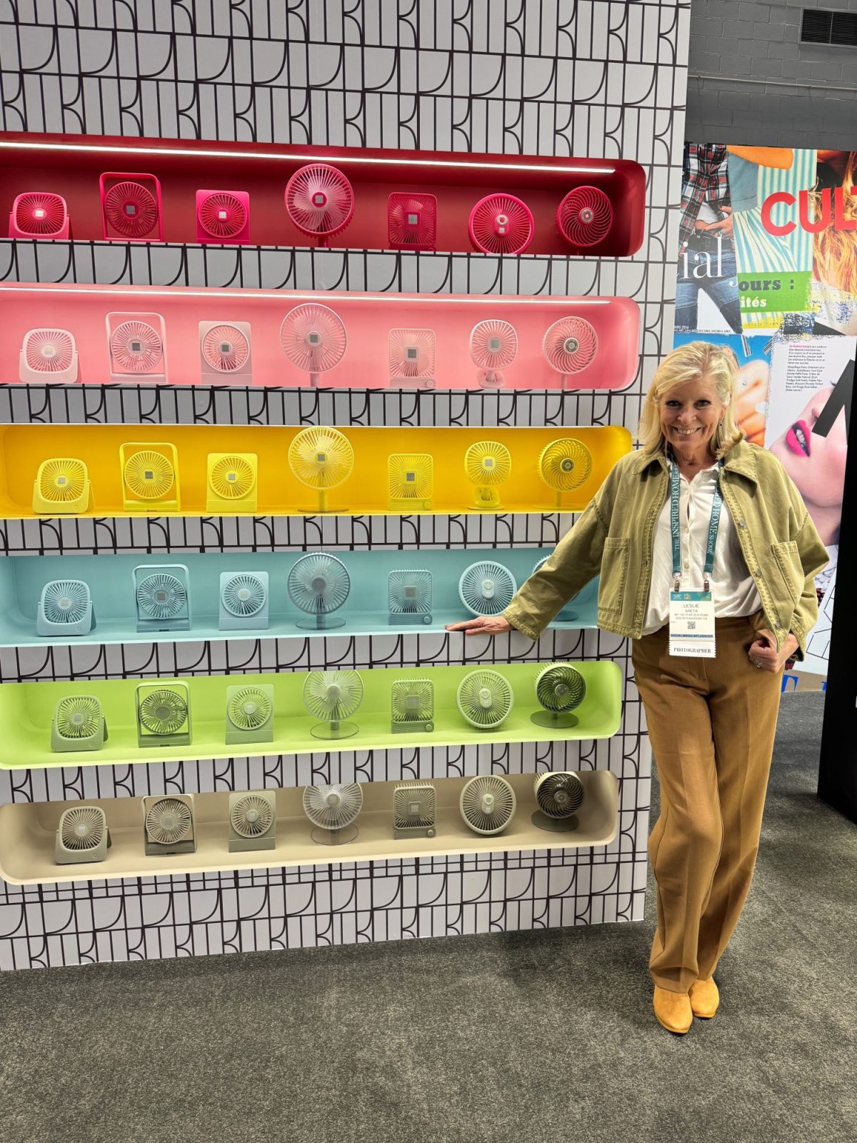 A woman stands beside a wall display of colorful, circular fans arranged in rows. She is smiling and pointing at a beige fan.