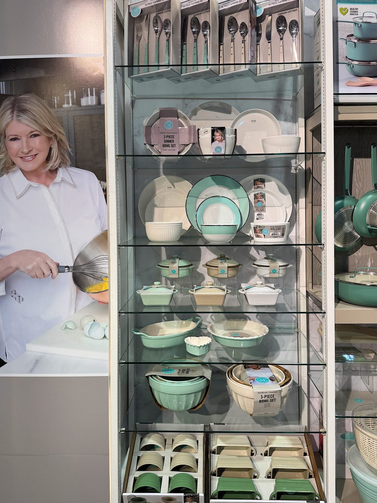 Display of kitchenware, including pots, bowls, and utensils, in white and teal. Packaging visible on shelves. Poster of a person cooking in the background.
