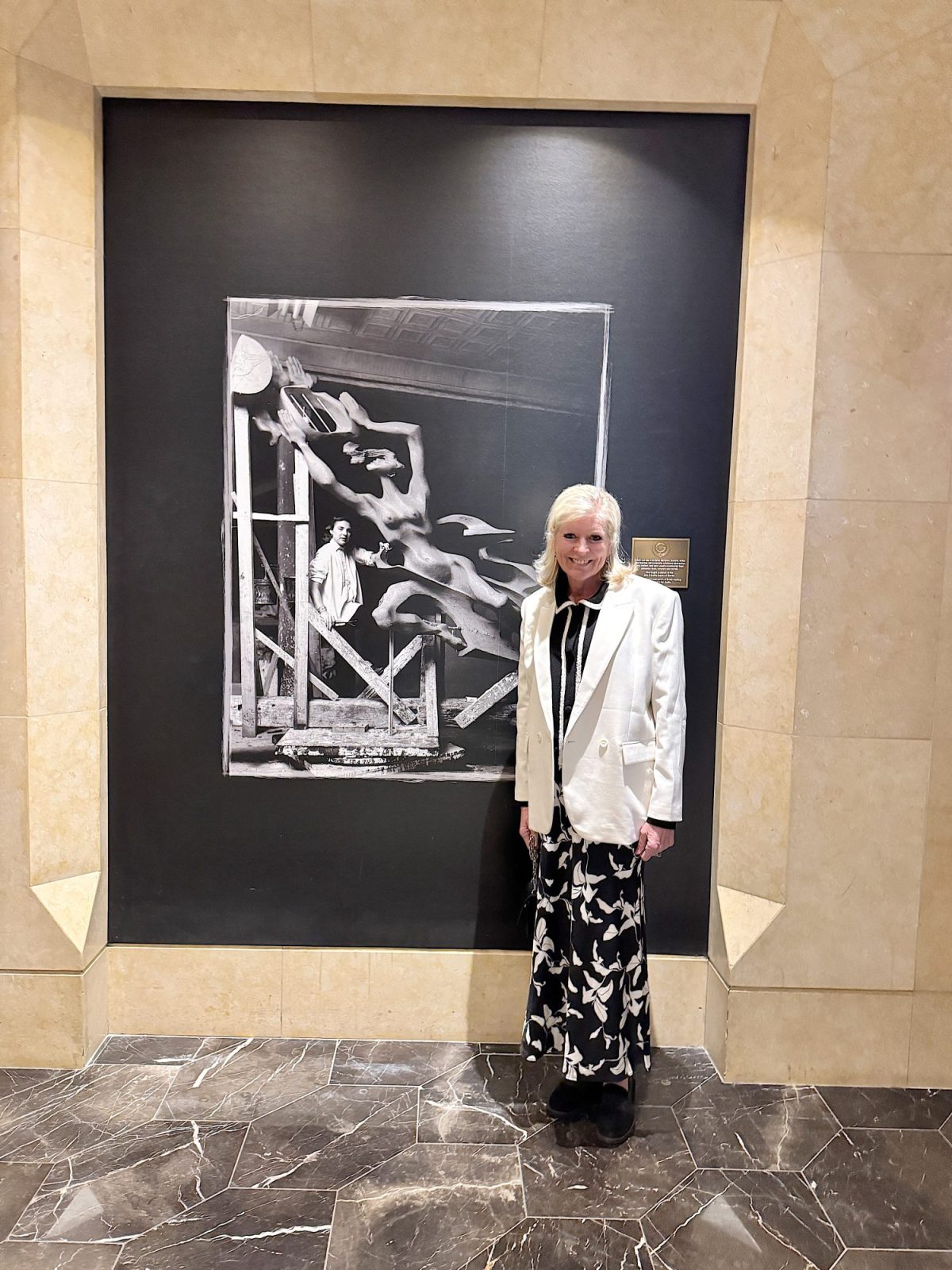 Person in a white blazer and floral skirt standing next to a large, monochrome framed artwork in a marble-tiled hallway.