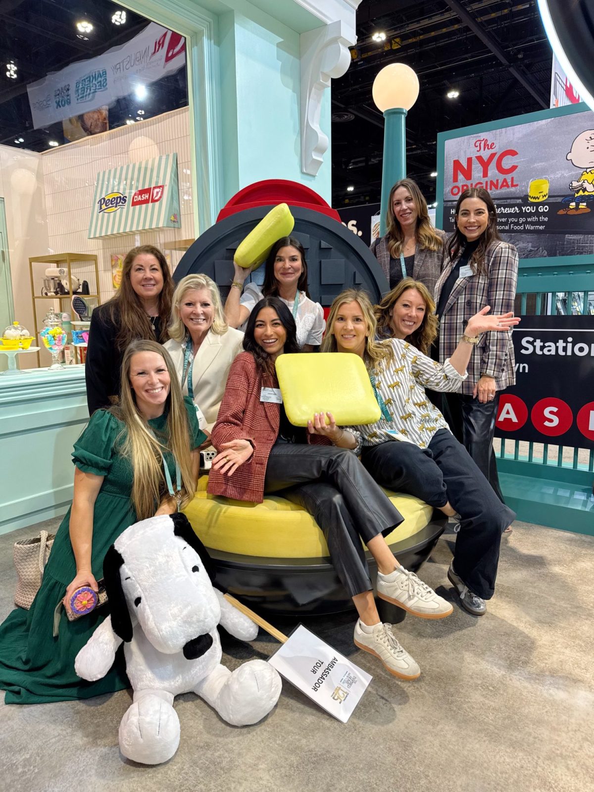 A group of nine women posing around a large Pez dispenser display, some seated, others standing. A Snoopy toy sits in the foreground. The setting looks like an exhibition space.