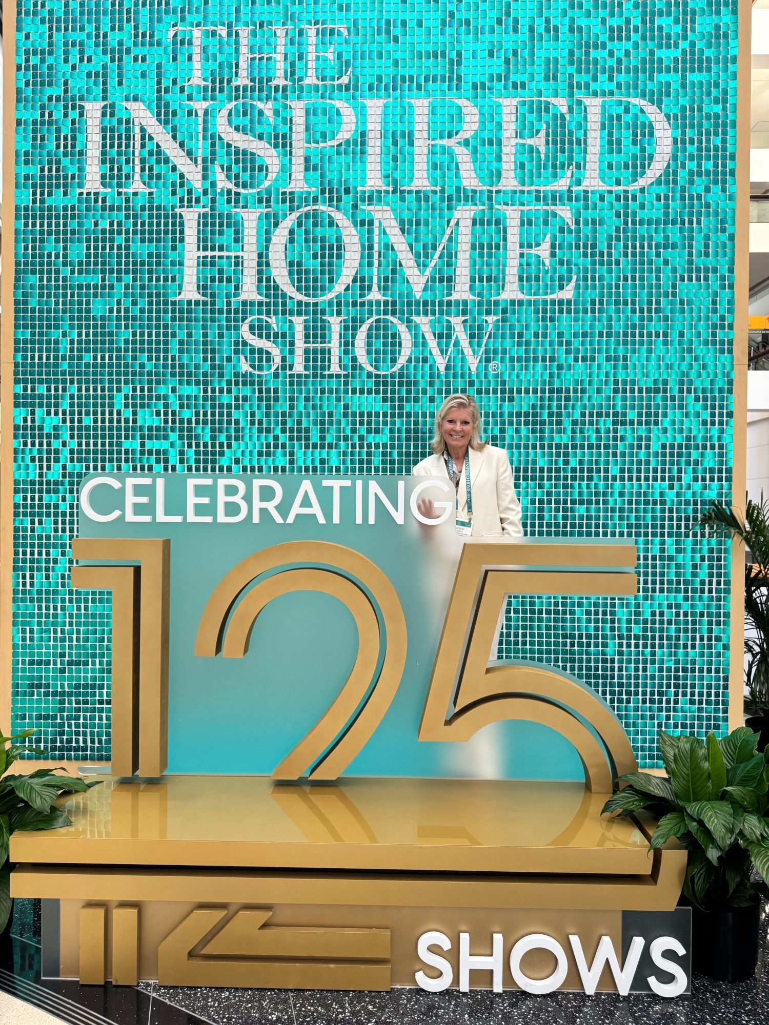 Person standing behind a large sign reading "Celebrating 125 Shows" at The Inspired Home Show in front of a turquoise-tiled background.