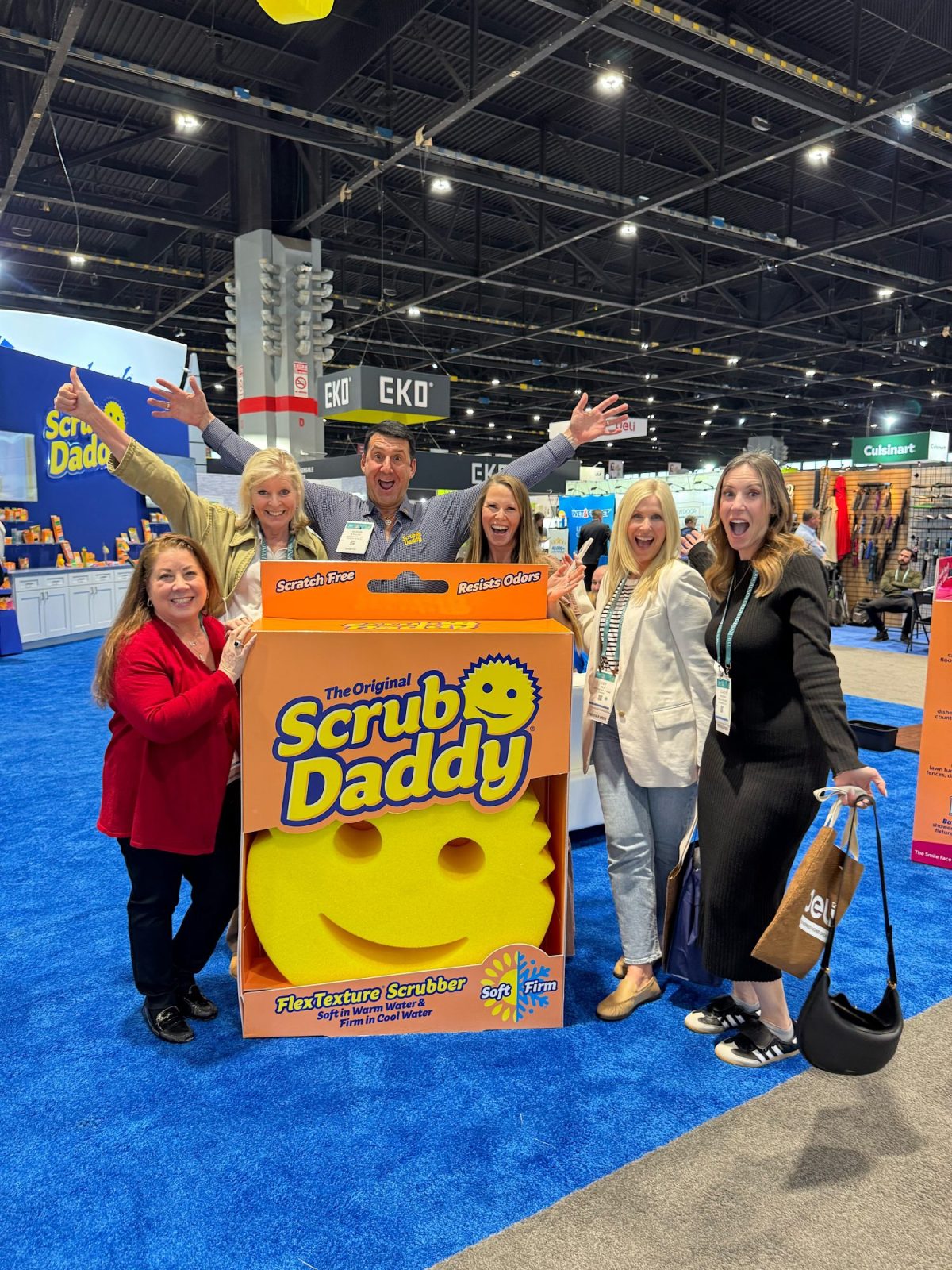A group of people pose with a large Scrub Daddy display box in a convention center. They appear cheerful and excited.