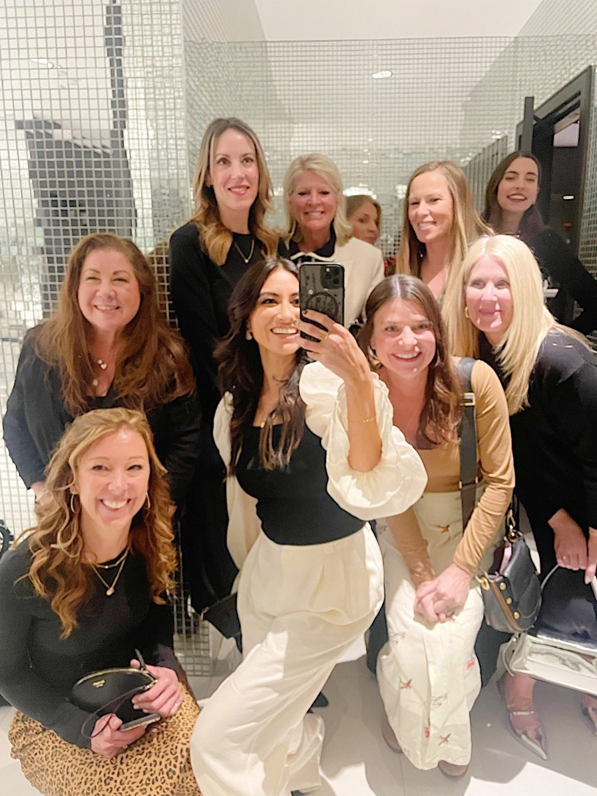 A group of nine women pose for a selfie in a mirrored room. They are dressed in casual to semi-formal attire and are smiling at the camera.