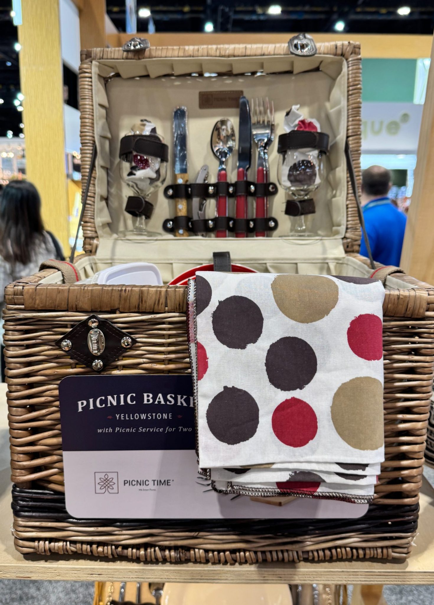 Wicker picnic basket with cutlery, glasses, and napkins inside. A polka dot napkin is draped over the side.