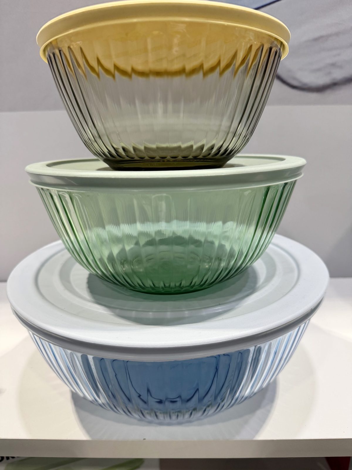 Three stacked glass bowls in yellow, green, and blue shades, each with a matching lid, displayed on a white surface.