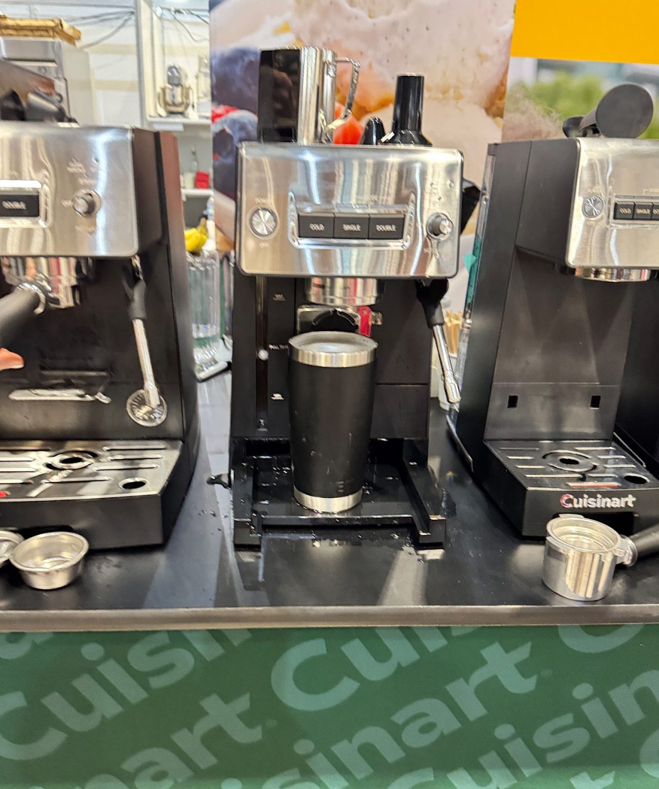 Silver and black espresso machine brewing into a black tumbler, surrounded by other coffee machines and accessories on a counter.