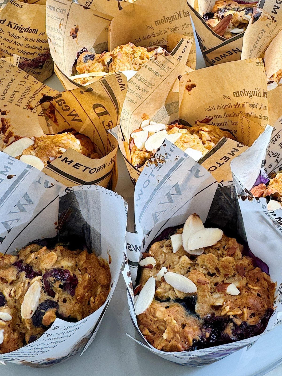 Multiple muffins wrapped in newspaper-themed paper, topped with almond slices and blueberries.