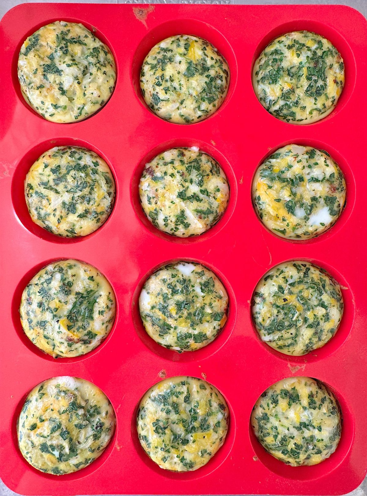 A red silicone muffin tray holds twelve baked vegetable egg muffins, featuring visible bits of green herbs.