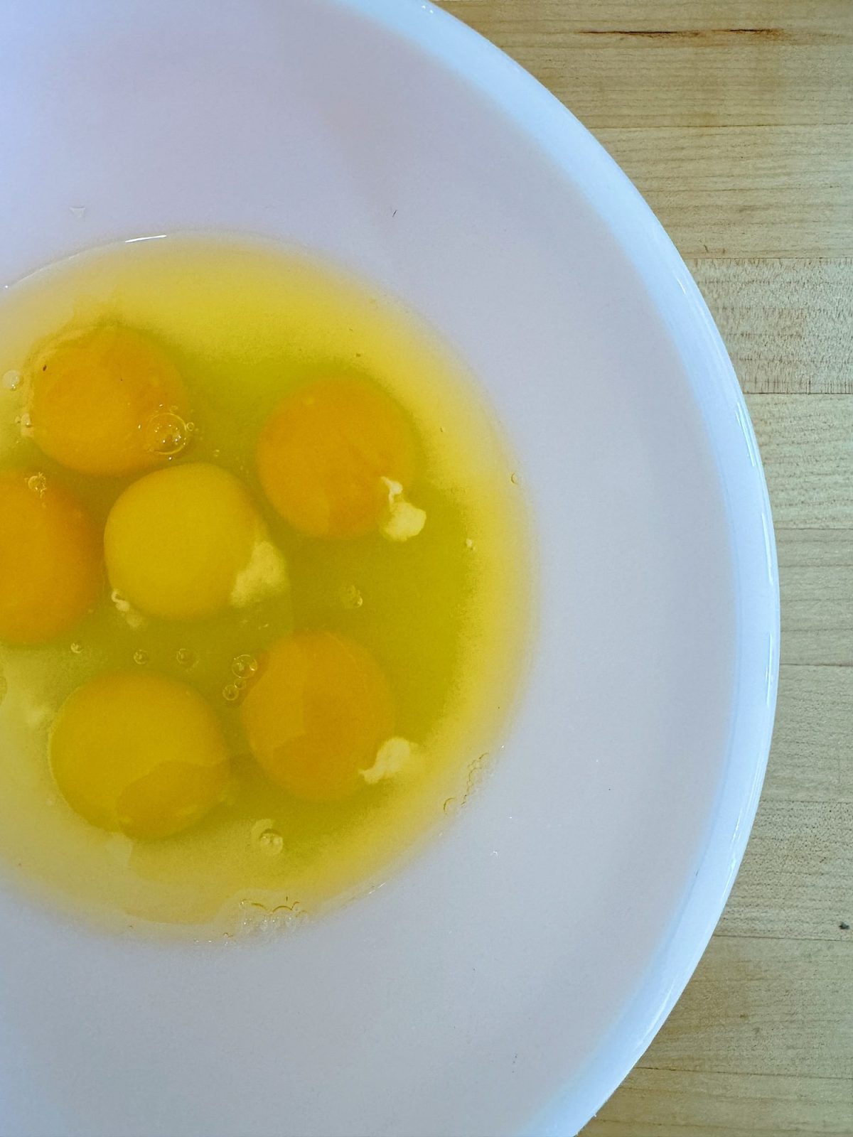 Six raw eggs cracked into a white bowl on a wooden surface.