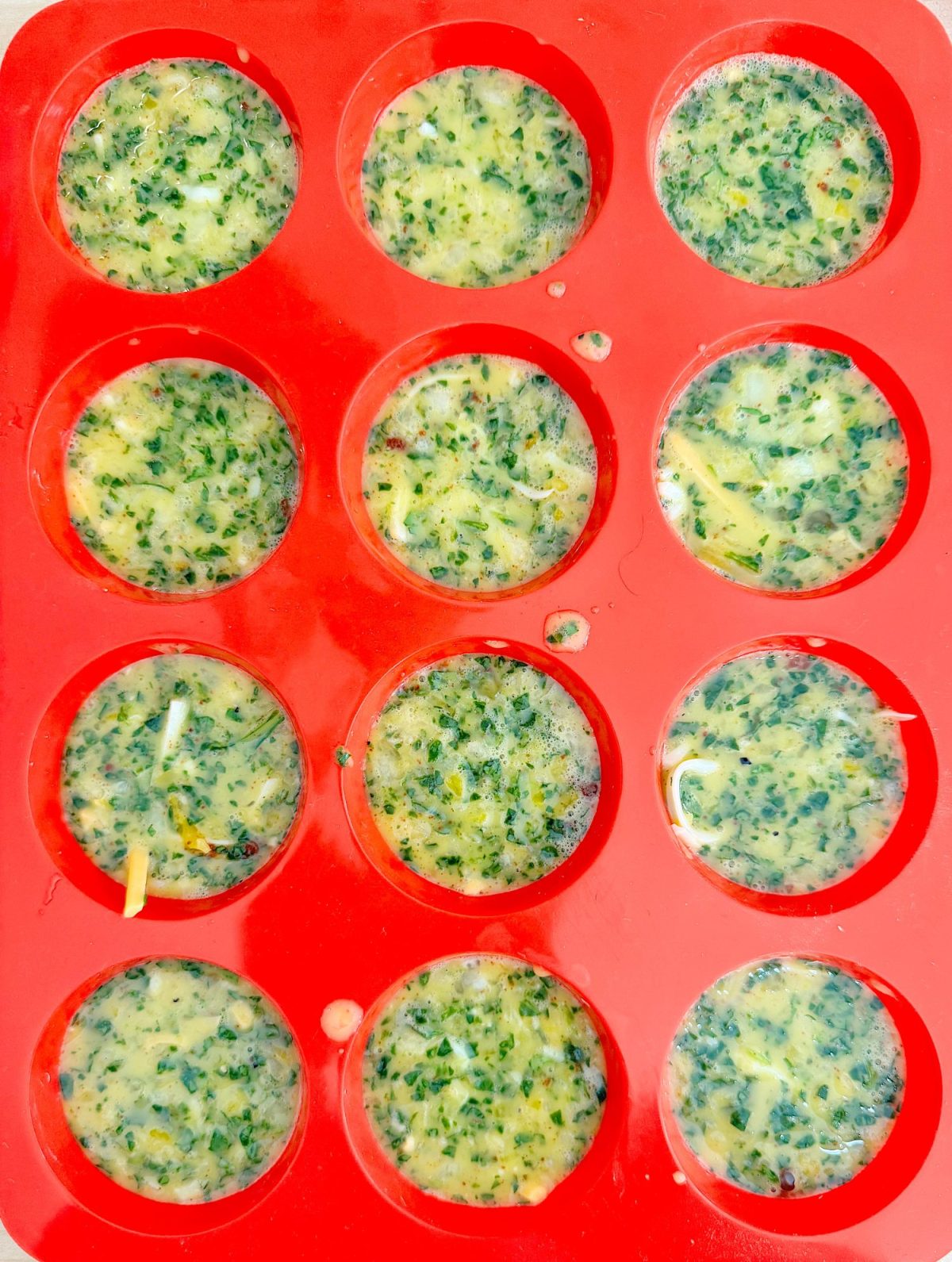 Red silicone mold filled with twelve portions of a green and yellow mixture, likely herbs and cheese, prepared for freezing or baking.