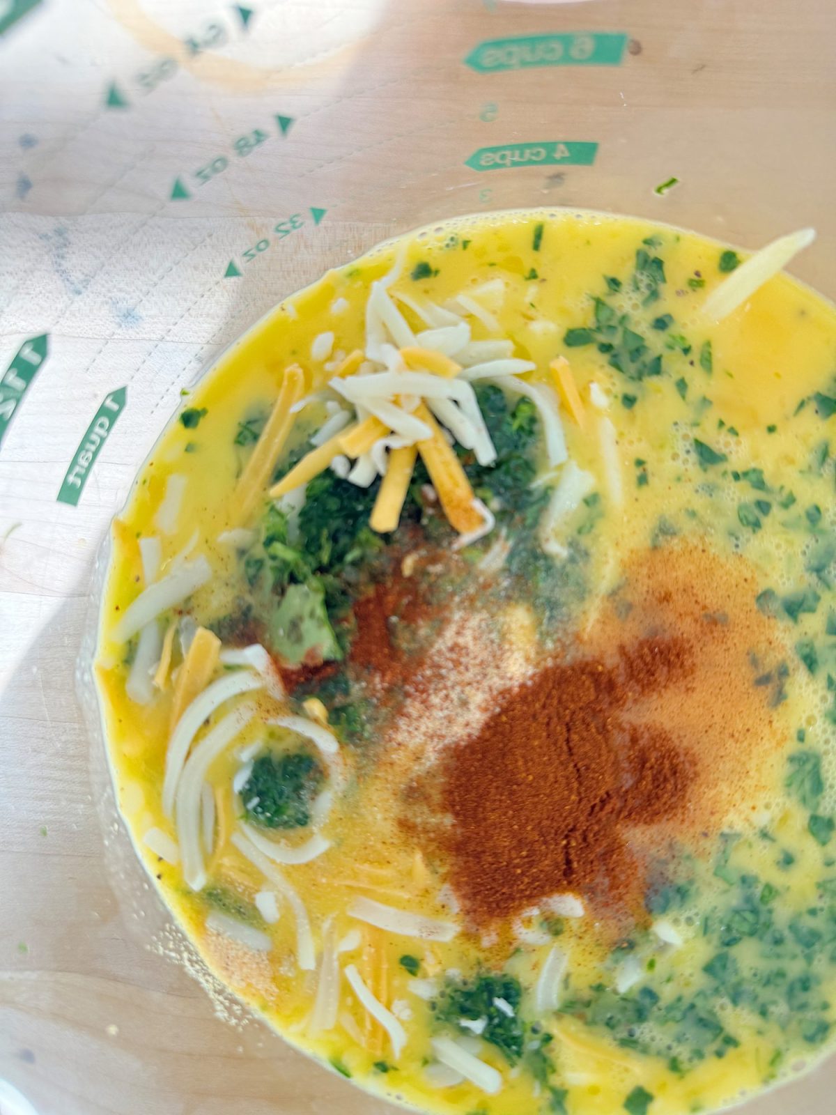 A bowl with beaten eggs, shredded cheese, green herbs, and paprika on top.