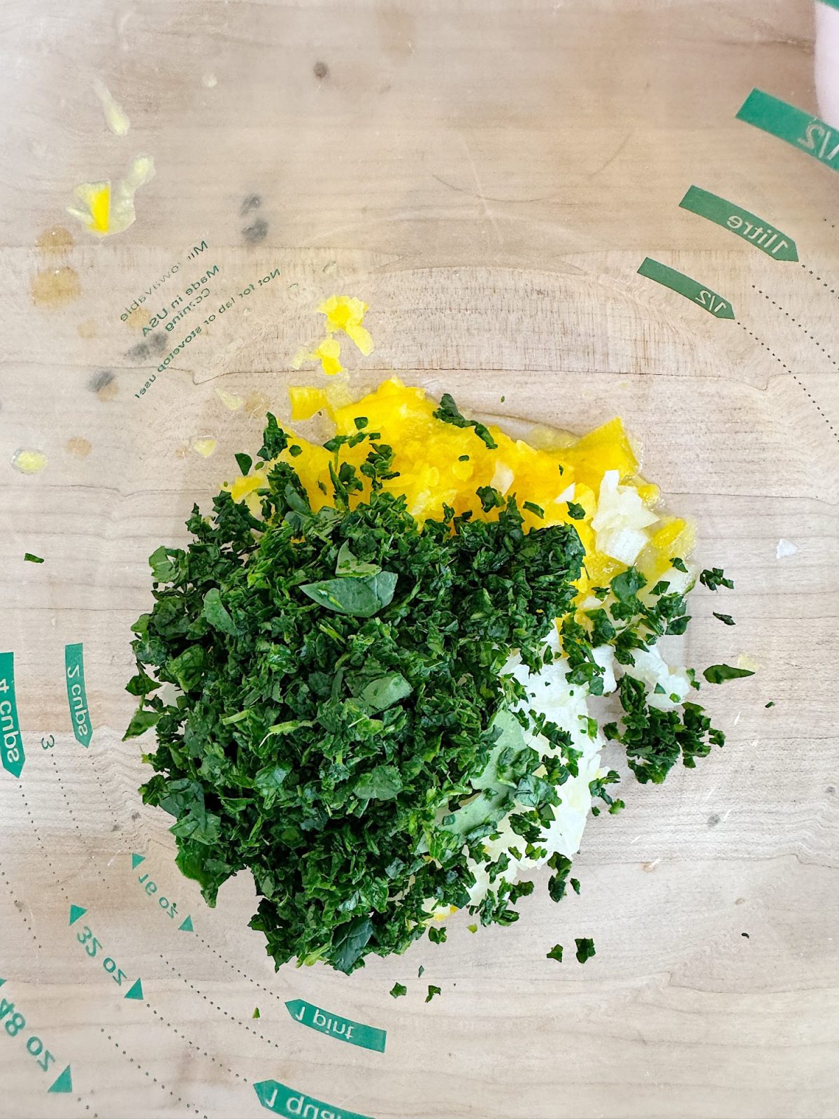 Chopped greens, diced yellow peppers, and grated cheese on a clear circular mat with measurement markings on a wooden surface.