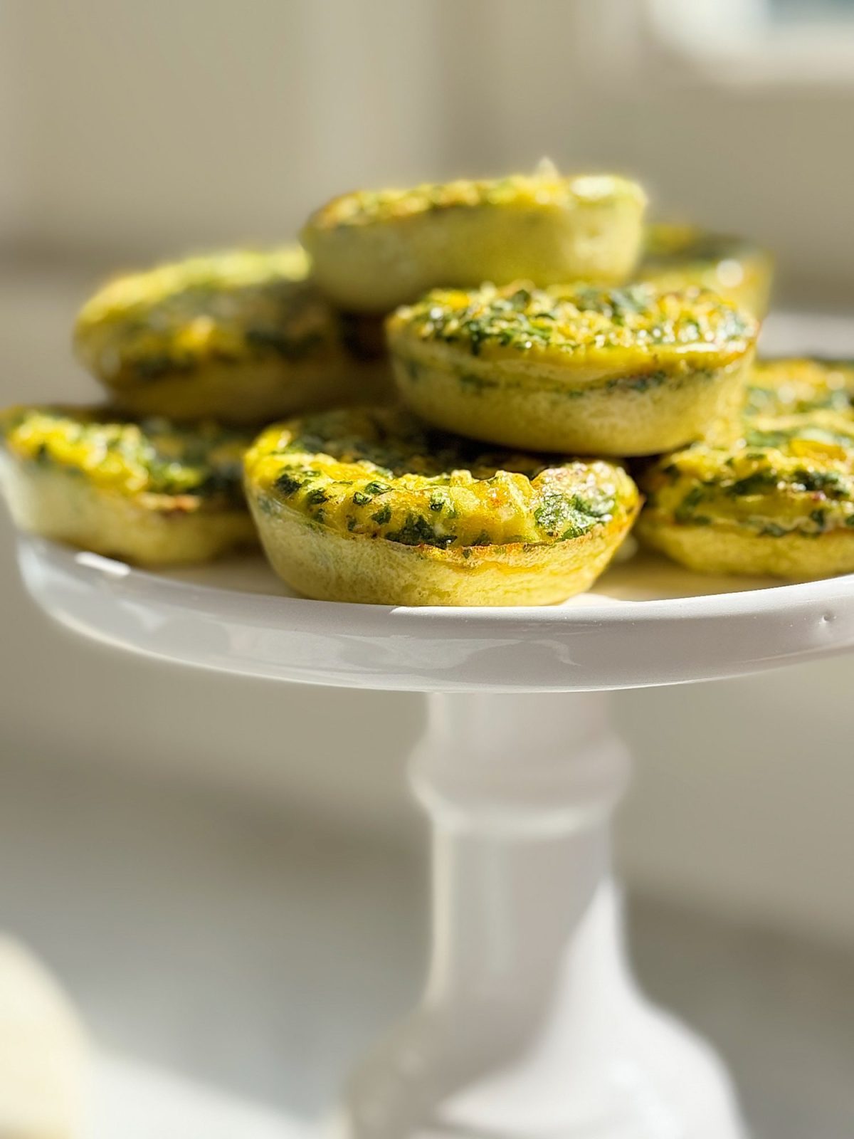 Mini spinach and cheese quiches stacked on a white pedestal cake stand in soft natural light.