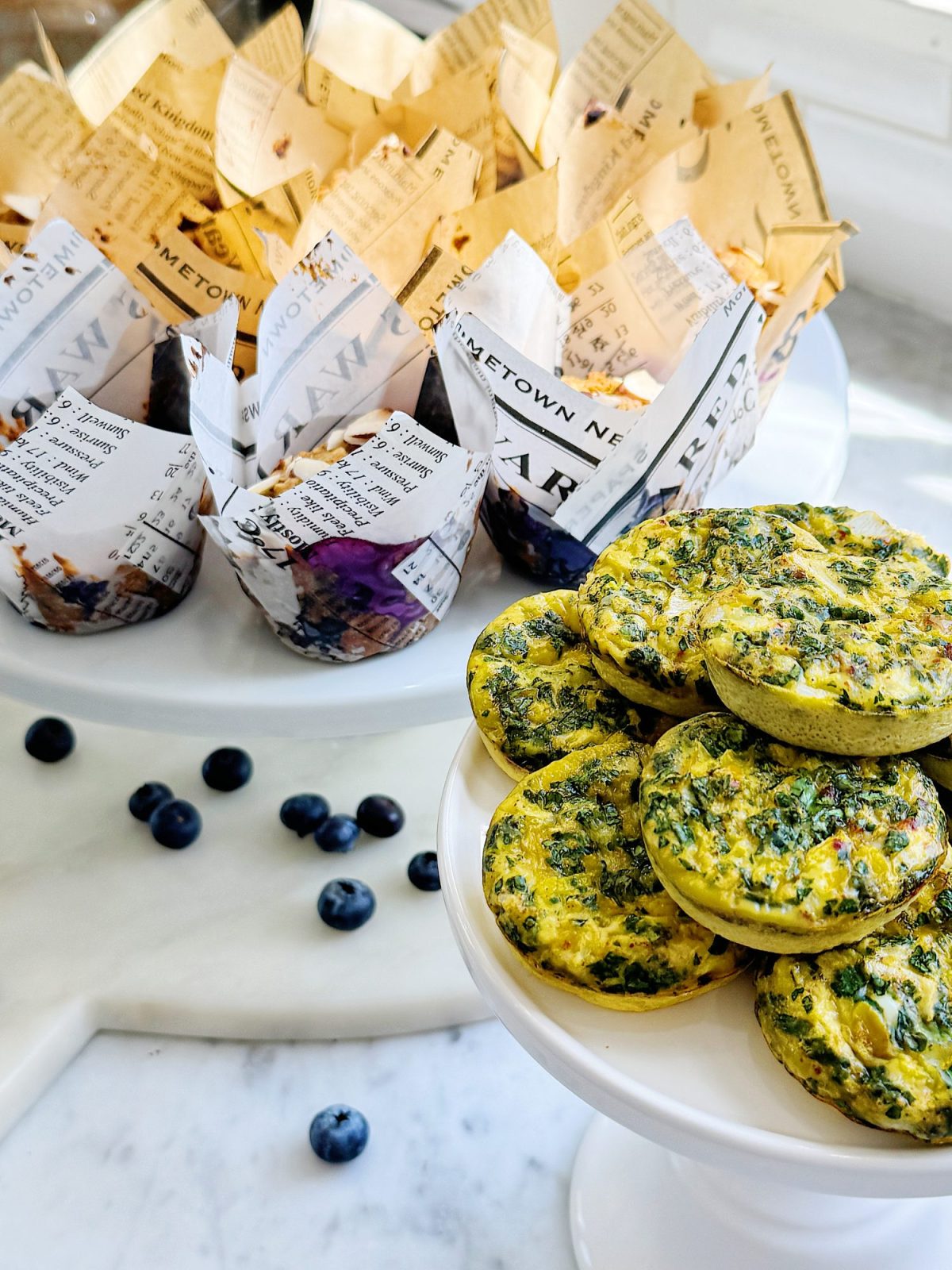 Muffins in newspaper wrappers on a cake stand, spinach and cheese frittatas on a plate, and scattered blueberries on a marble surface.
