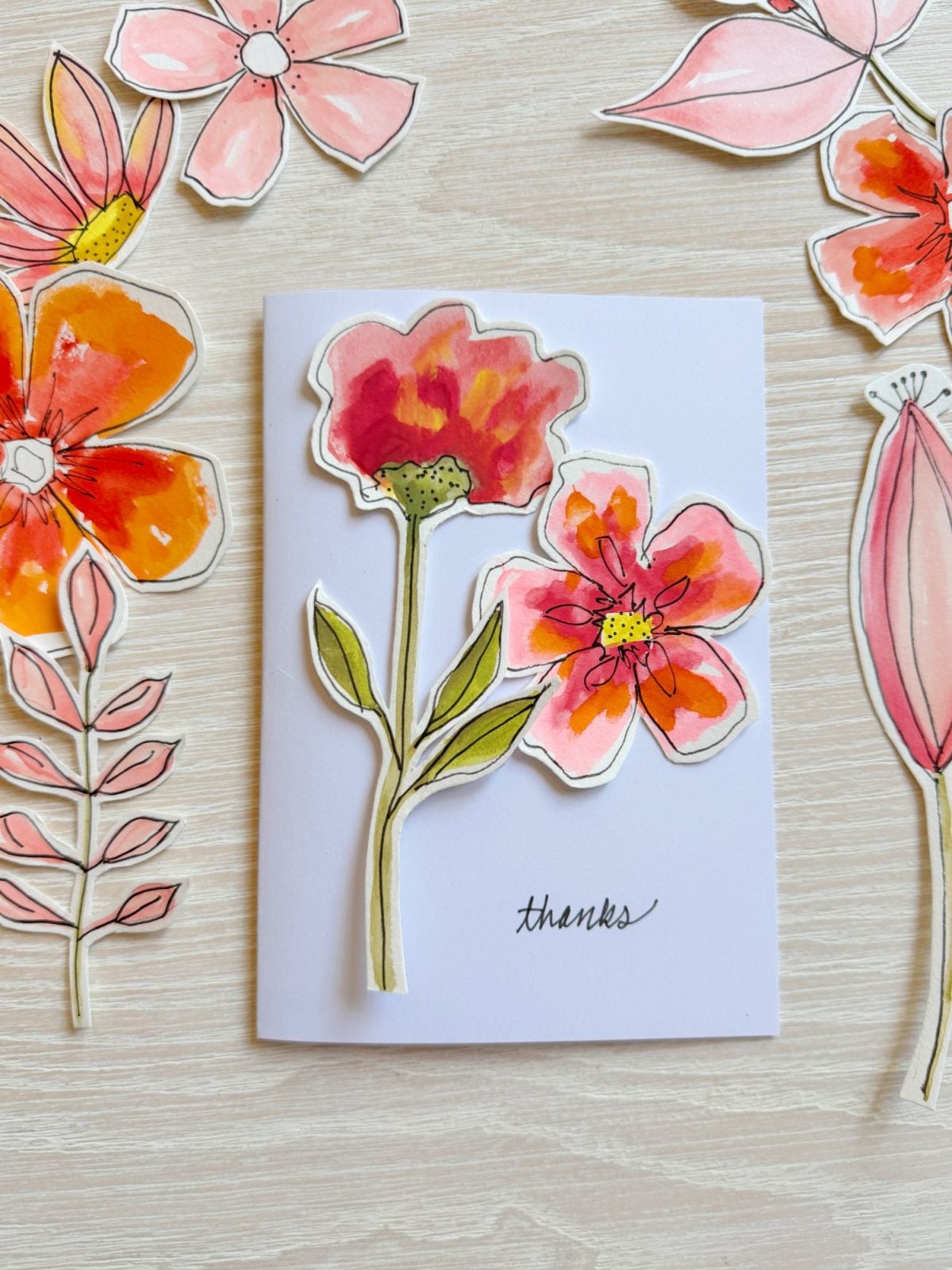 A white card with illustrated red and pink flowers and the word "thanks," surrounded by more floral cutouts on a light wood surface.