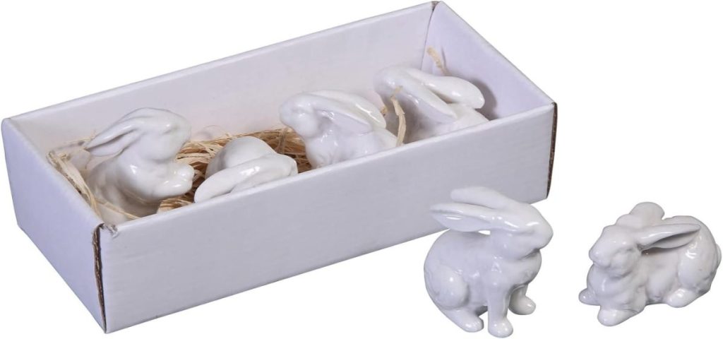 Four white ceramic rabbit figurines in a white cardboard box with straw filling, accompanied by Easter Eggs and two more rabbit figurines placed outside the box.