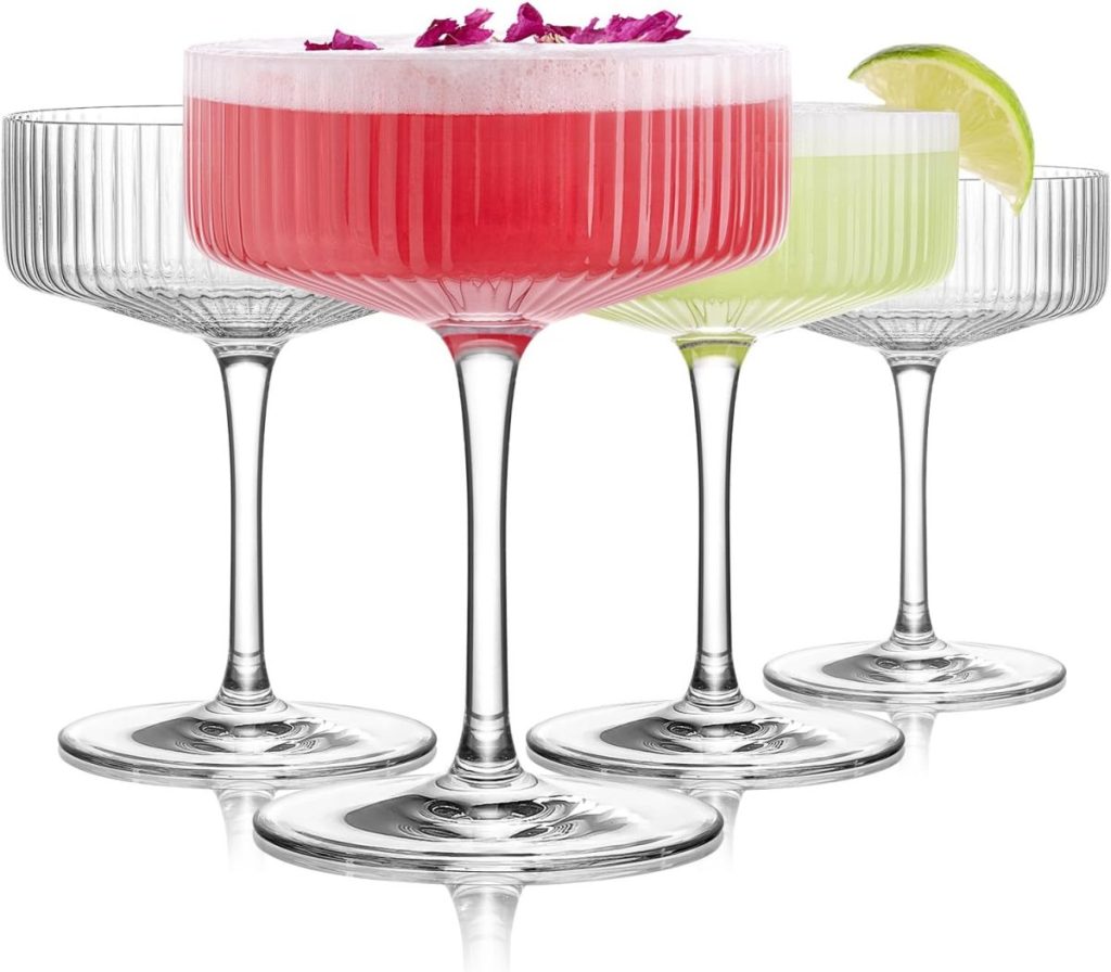 Four ribbed coupe glasses, two empty and two filled with cocktails; one red drink garnished with petals, one green lemon vodka cocktail garnished with a lime wedge.
