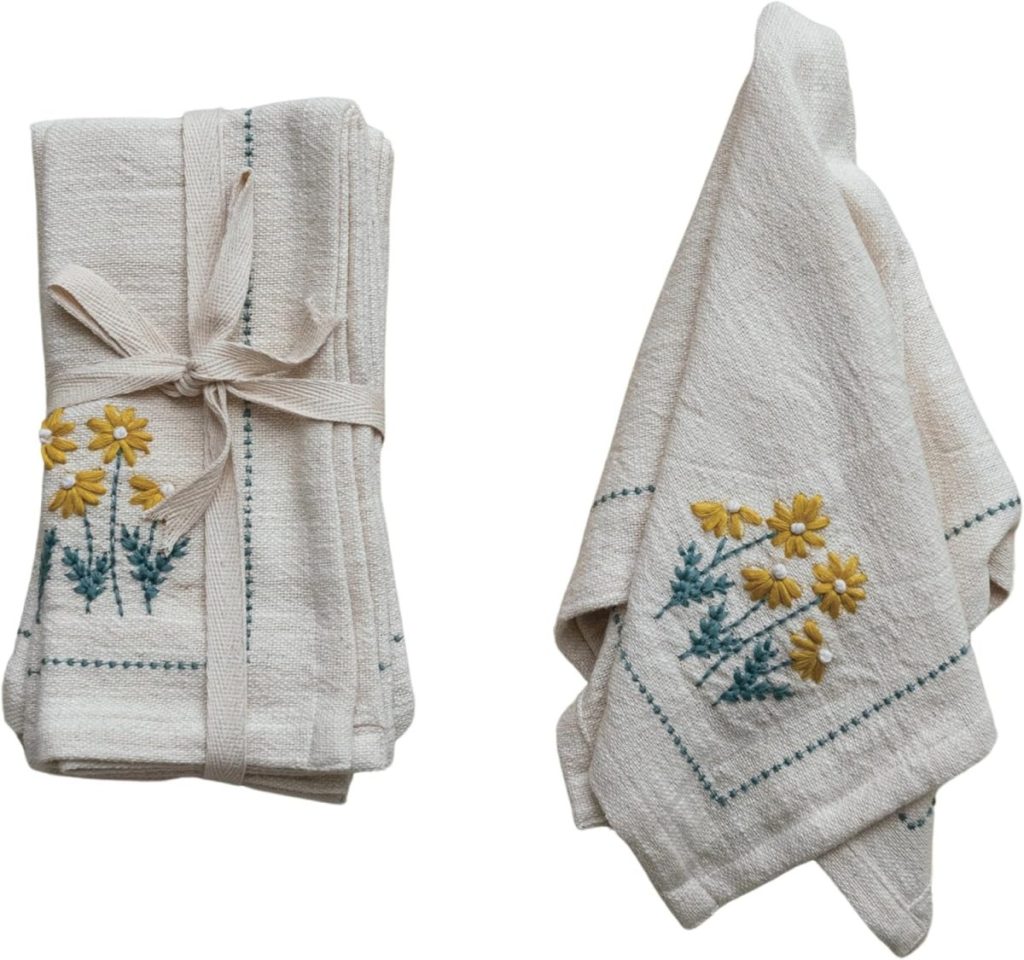 Two white cloth napkins with embroidered yellow flowers; one is folded and tied with a ribbon, the other is unfolded and draped—perfect for displaying alongside Easter eggs on your holiday table.