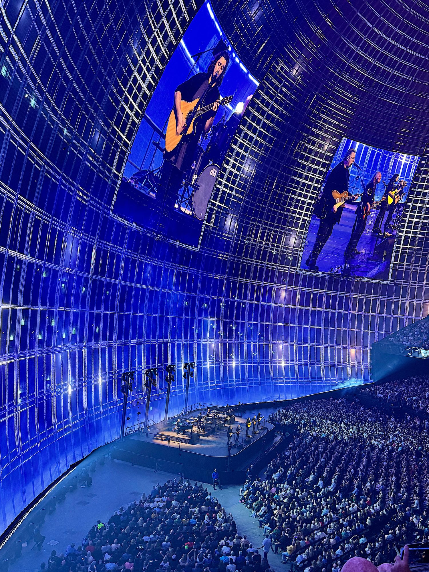 Large indoor concert in a futuristic venue with blue lighting. The stage features musicians playing guitars, with their images displayed on overhead screens. A large audience is seated below.