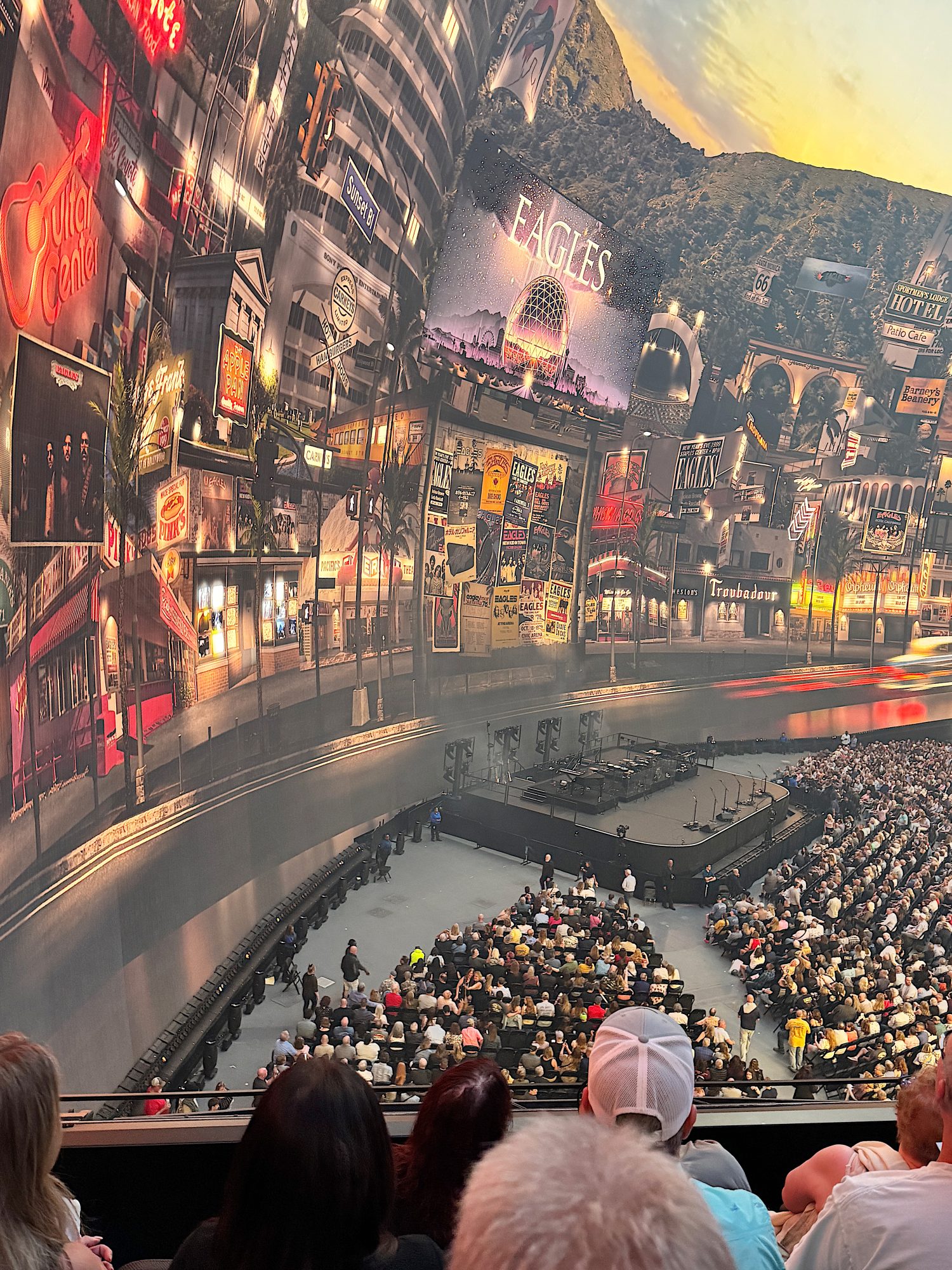 An audience sits before a stage in a theater, with a large, curved backdrop displaying various colorful billboards and signs.