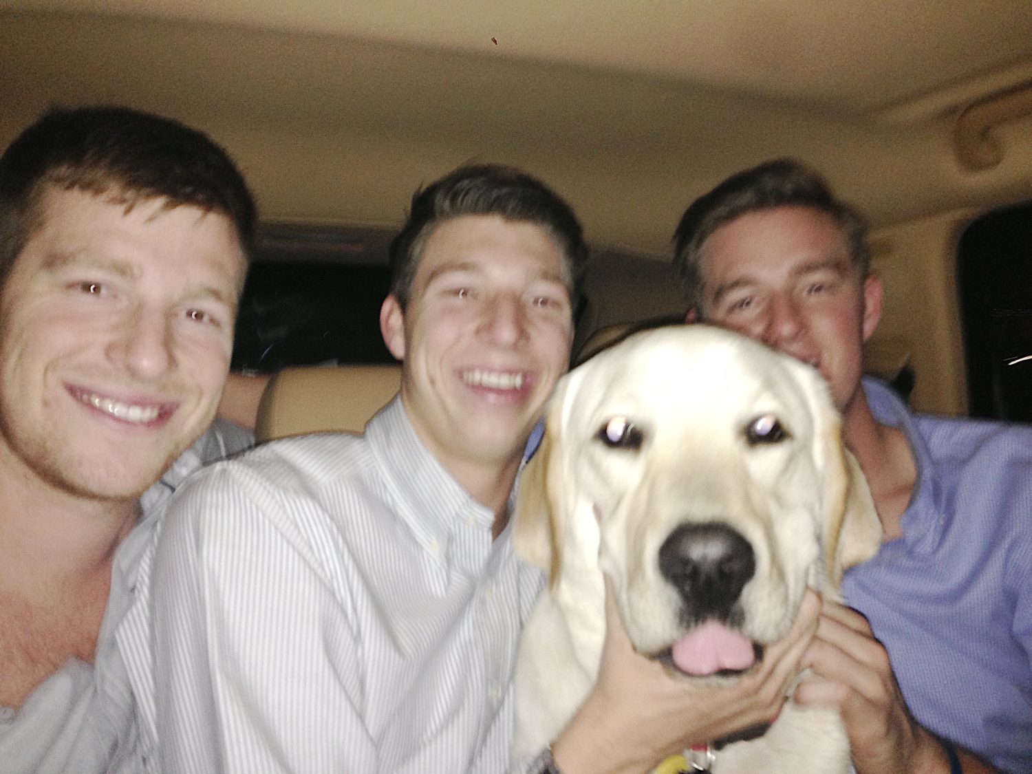 Three smiling people and a Labrador sitting together inside a car. The dog is in the center, looking at the camera with its tongue slightly out.