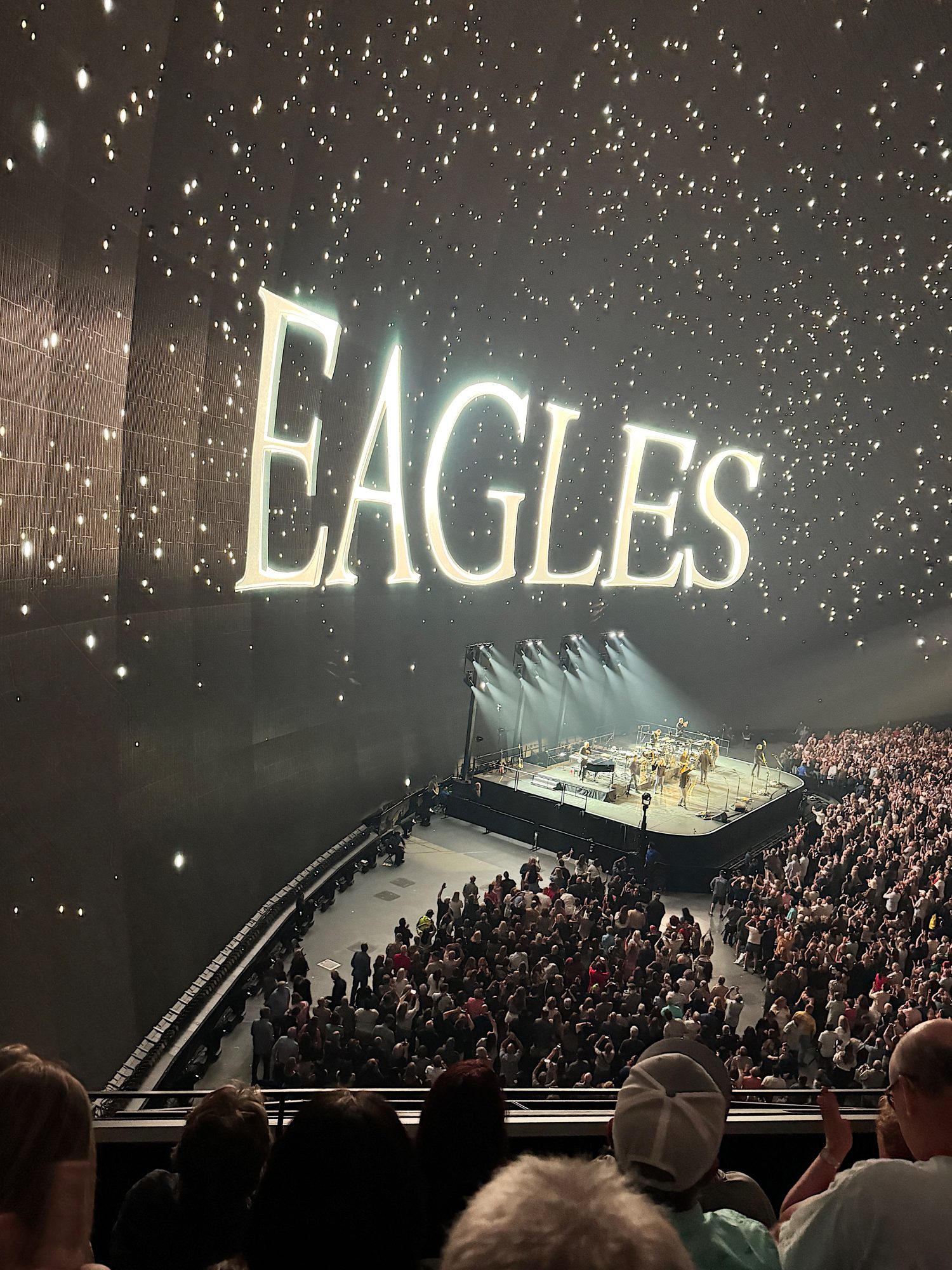 Concert stage with a large "Eagles" sign above. A band performs to a packed audience in a dimly lit venue with star-like lights.