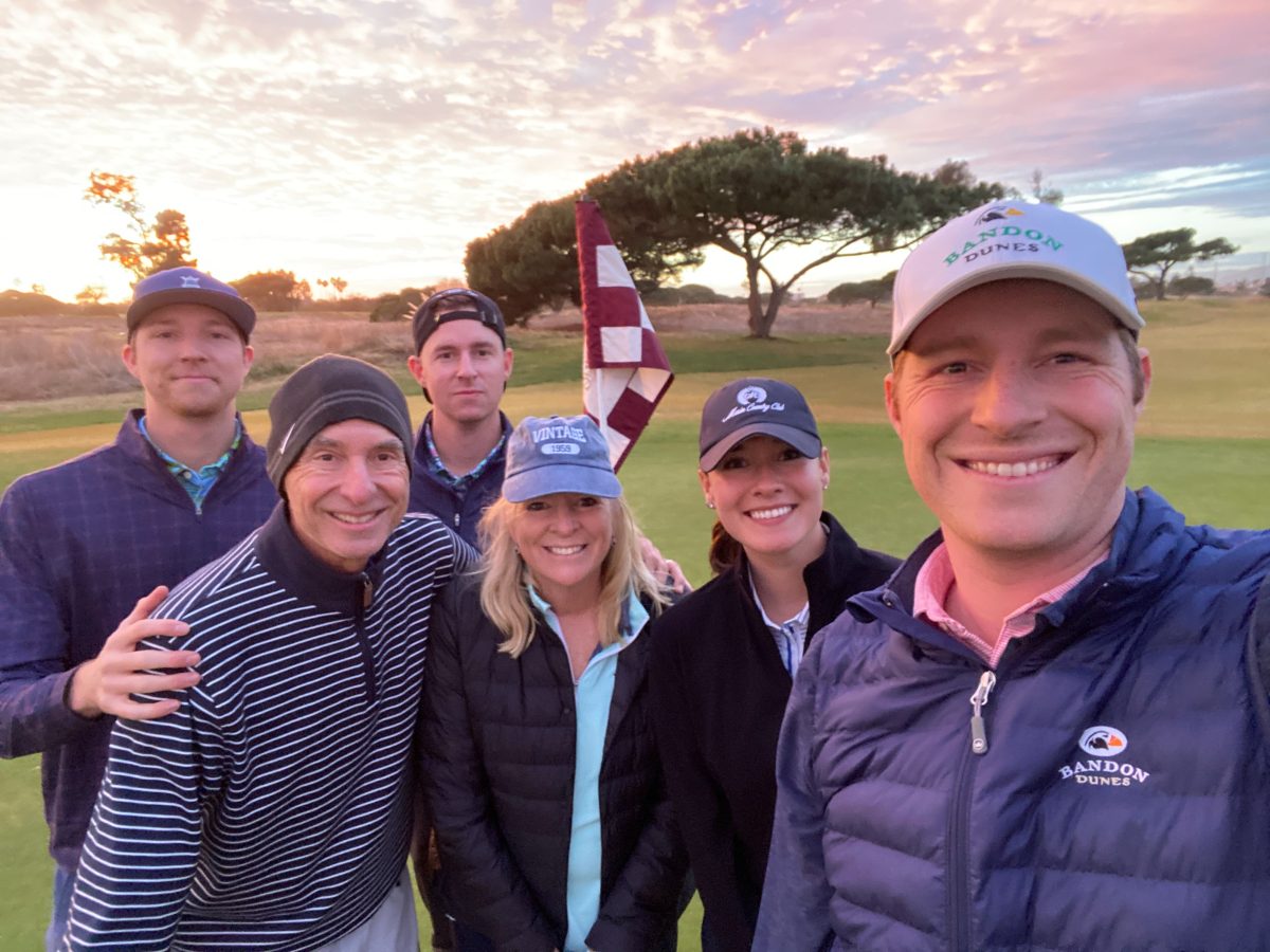 Six people pose for a group photo on a golf course at sunset, with a flagstick and trees visible in the background.