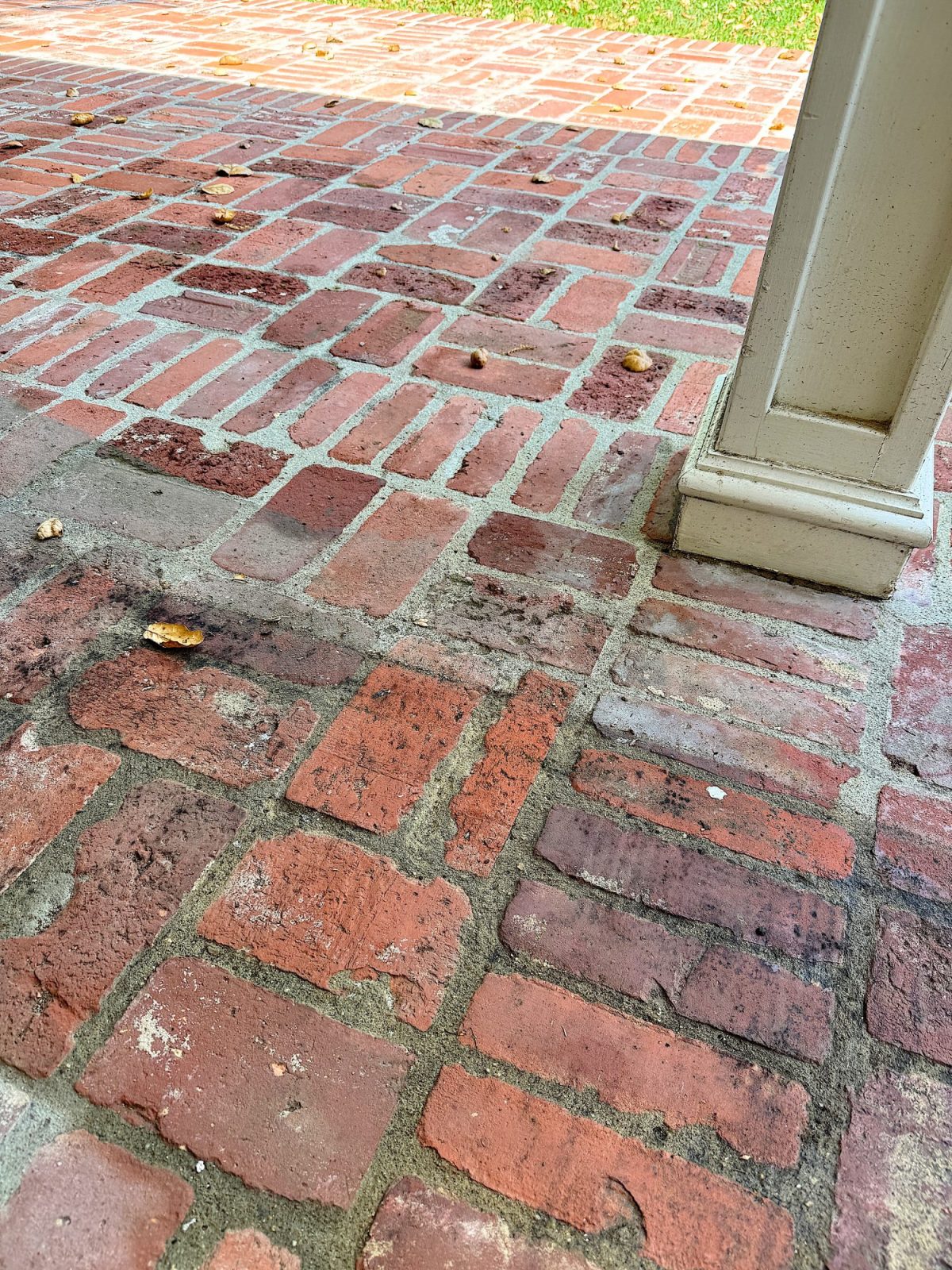 Red brick patio with scattered small debris and partial view of a white post, with grass visible in the background.