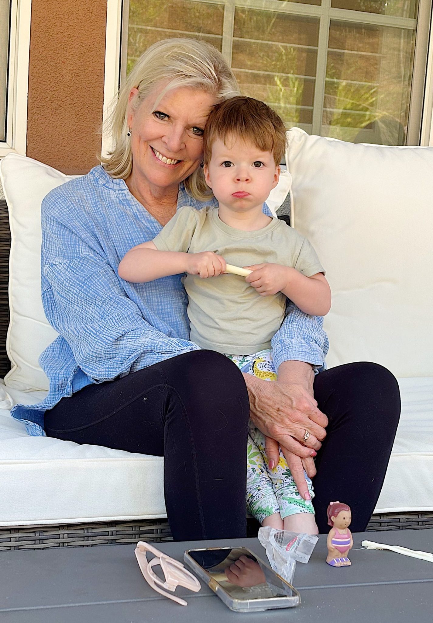 A woman and a child sit on a patio sofa. The child holds a popsicle stick. A phone, toy, and sunglasses are on the table in front of them.