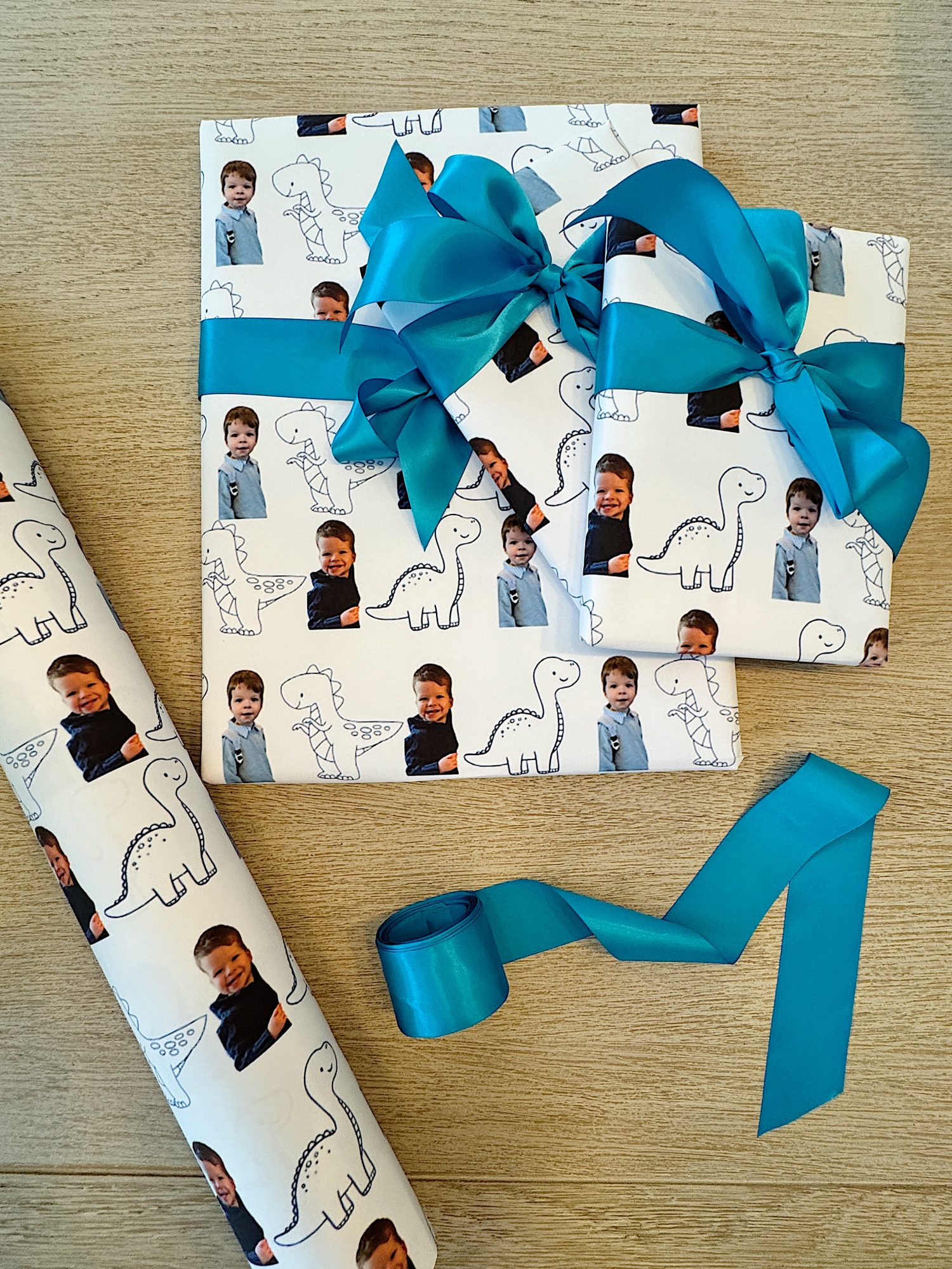 Three wrapped gifts with a custom photo and dinosaur pattern, tied with blue ribbons, next to a roll of the same wrapping paper and a loose ribbon.