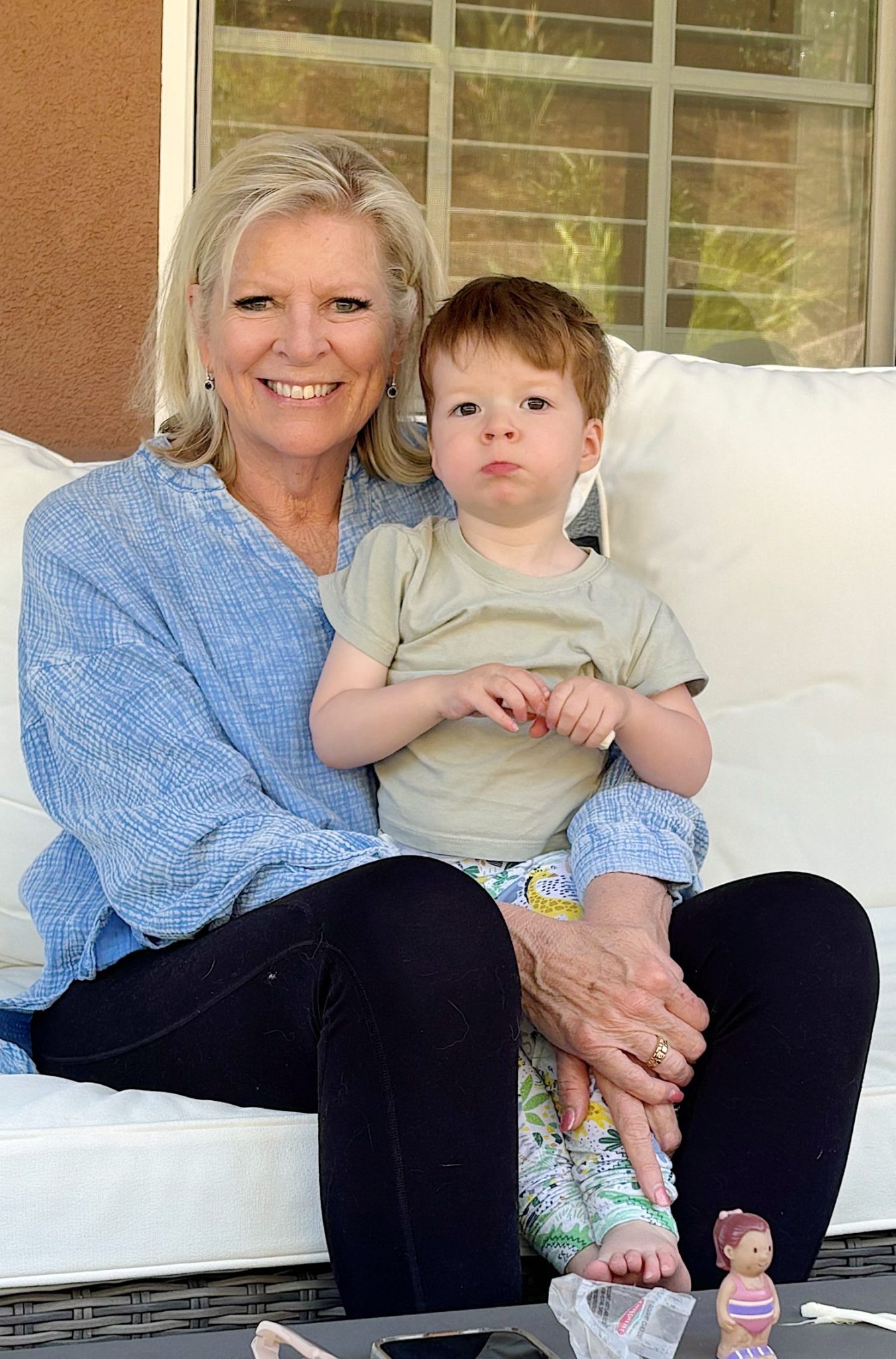 Leslie and a young child sitting on a white couch outdoors. The woman is smiling and wearing a blue shirt, while the child is looking forward and holding a toy.
