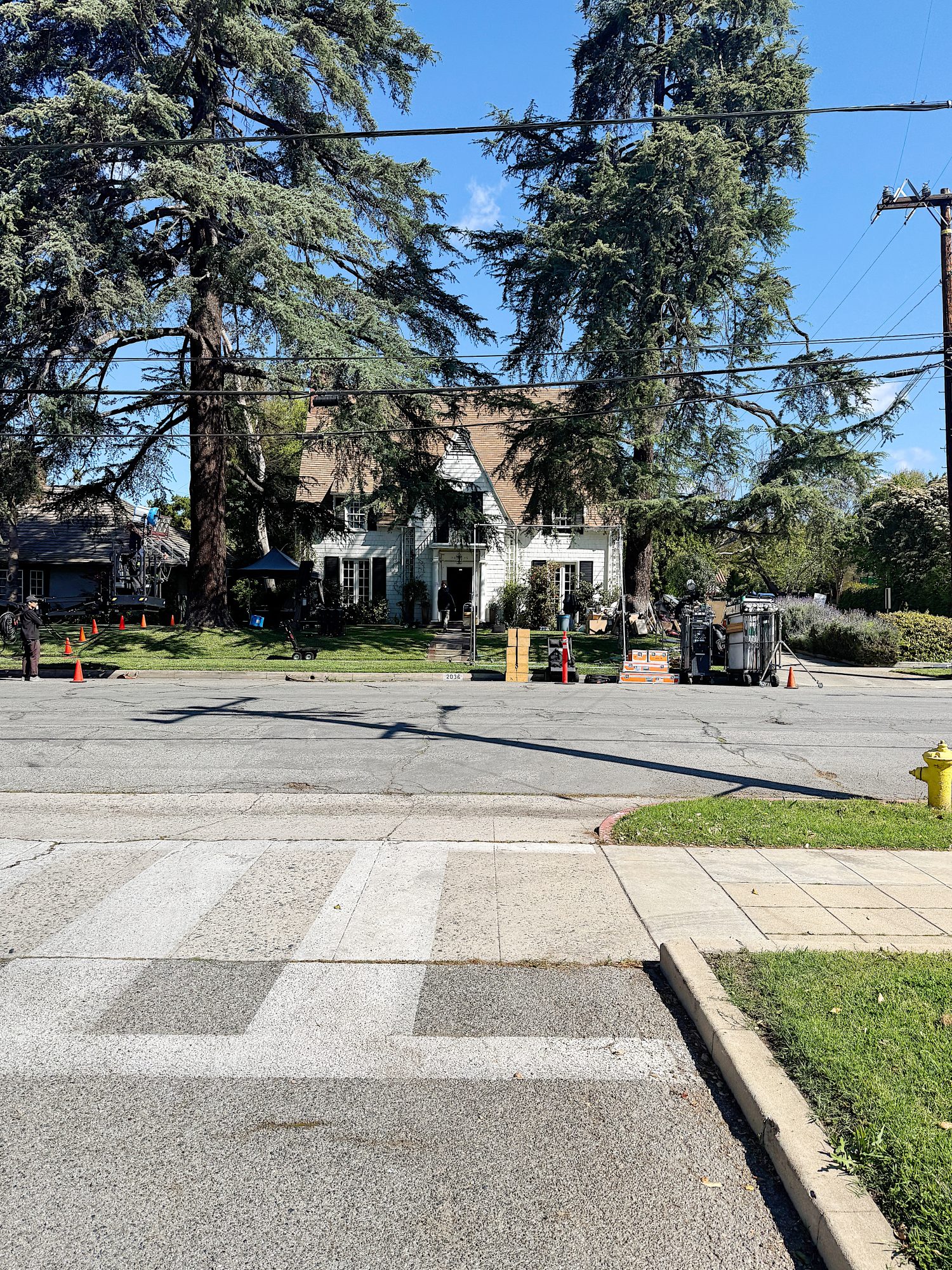 Residential street scene with a large house in the background, tall trees, utility wires, and orange traffic cones. A small crew is working on equipment nearby.