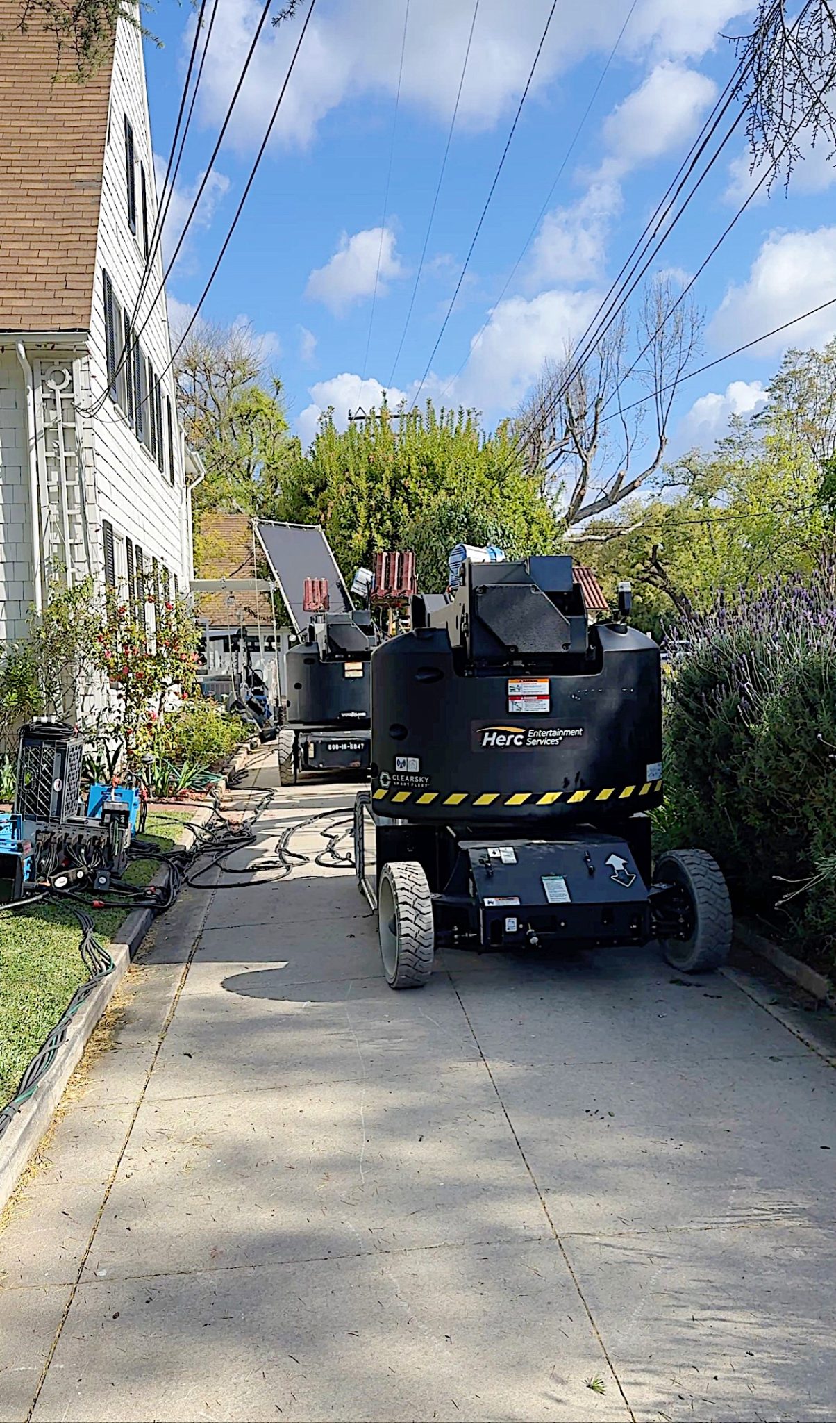 Two portable generators on a residential driveway, surrounded by cables and wires, with a white house on the side under a blue sky.