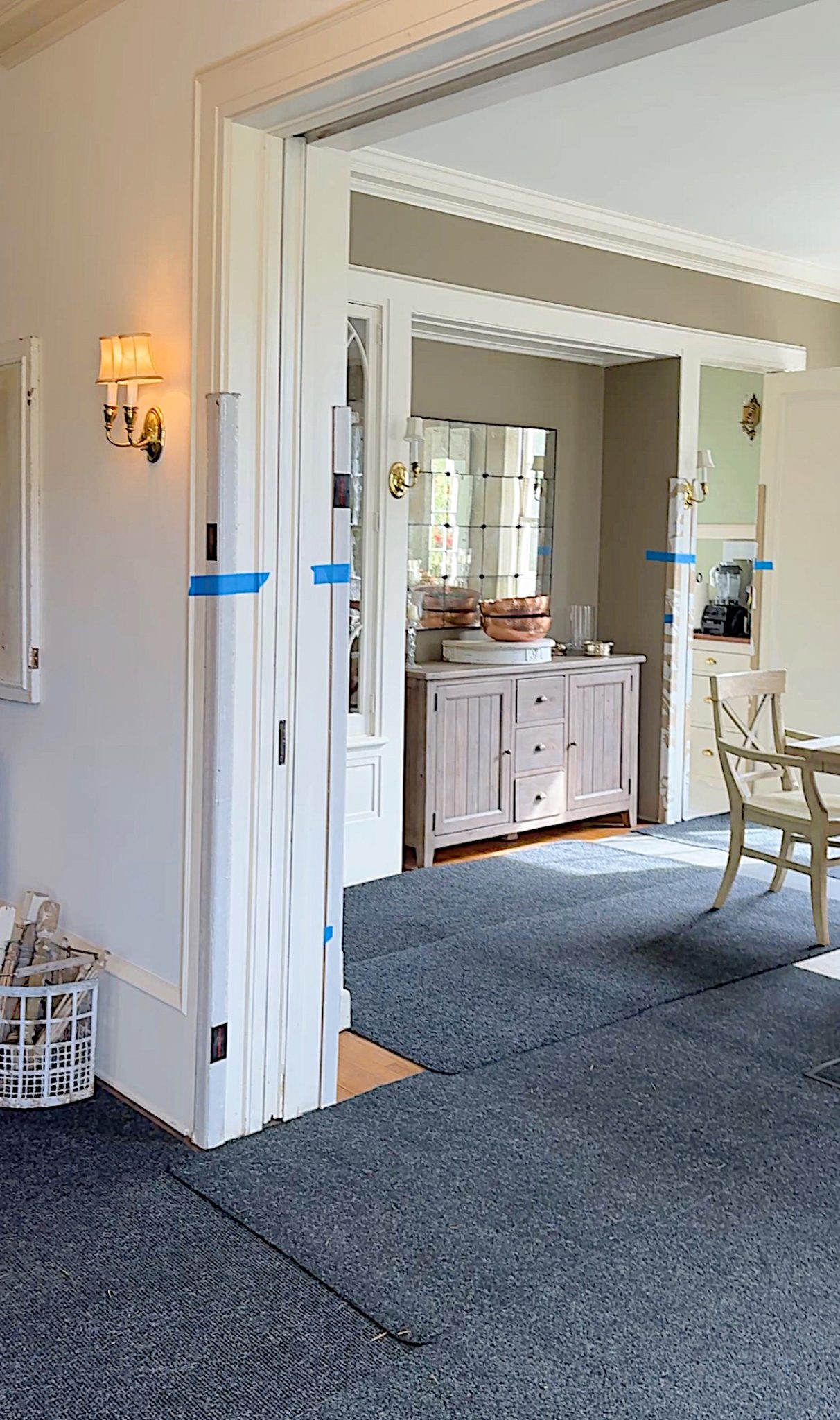 Interior view of a room with open double doors, a white sideboard with decorative items, and a dining table with chairs. The room has carpeted floors and is lit by wall sconces.