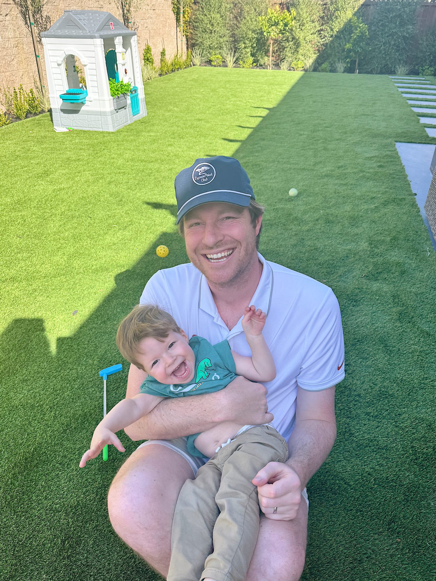Adult and child smiling while sitting on grass in a backyard with a small playhouse in the background.