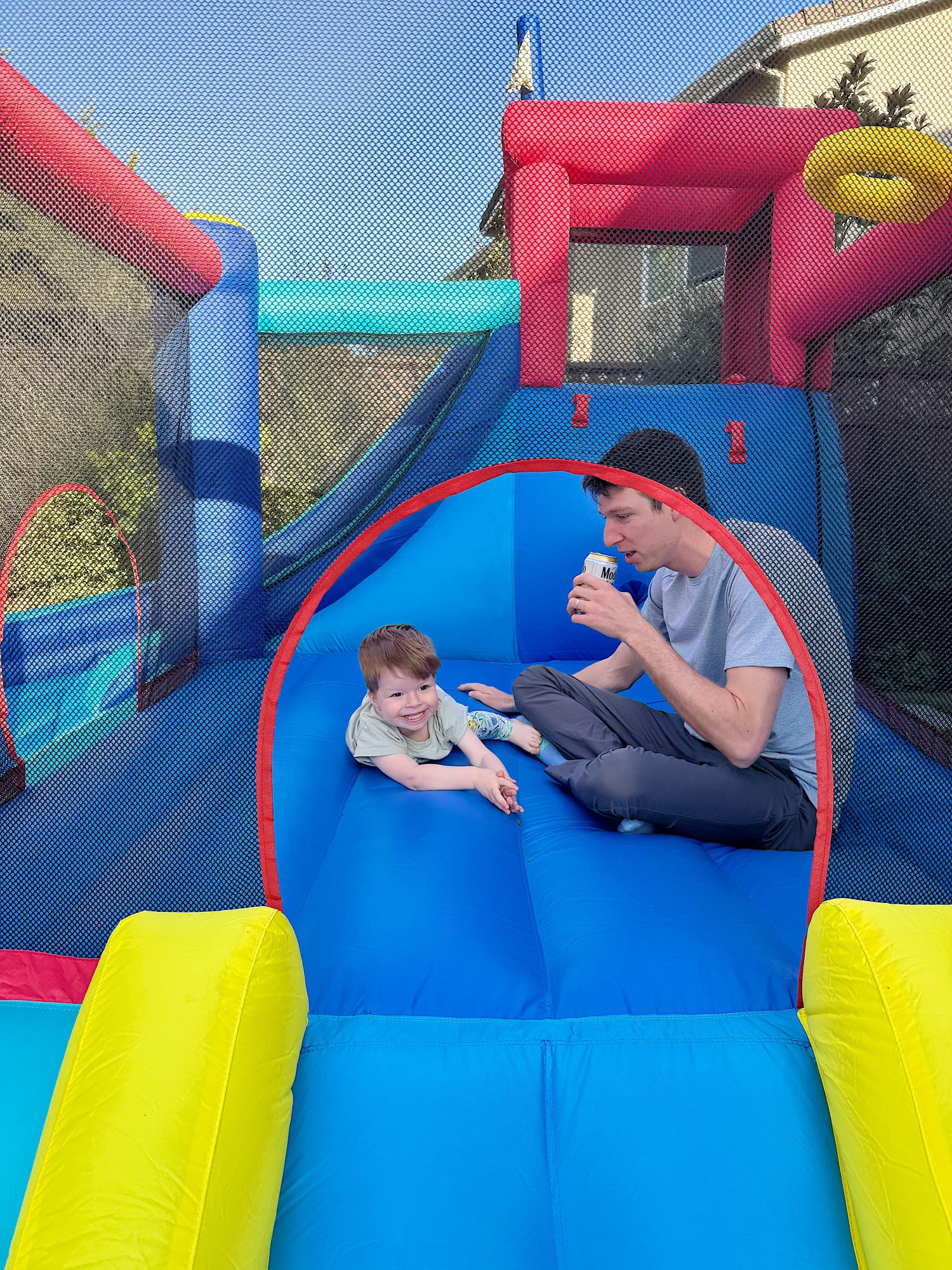 A child and an adult sitting inside a colorful inflatable bounce house; the adult is drinking from a cup.