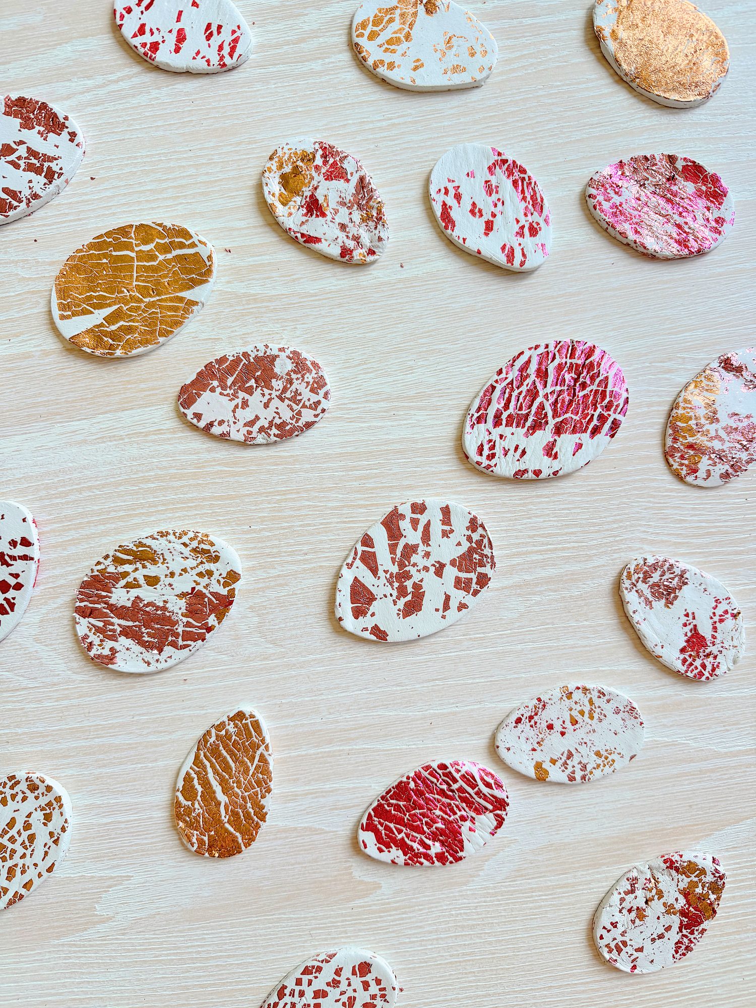 Oval pieces of textured material with red, gold, and copper patterns are scattered on a light wooden surface.