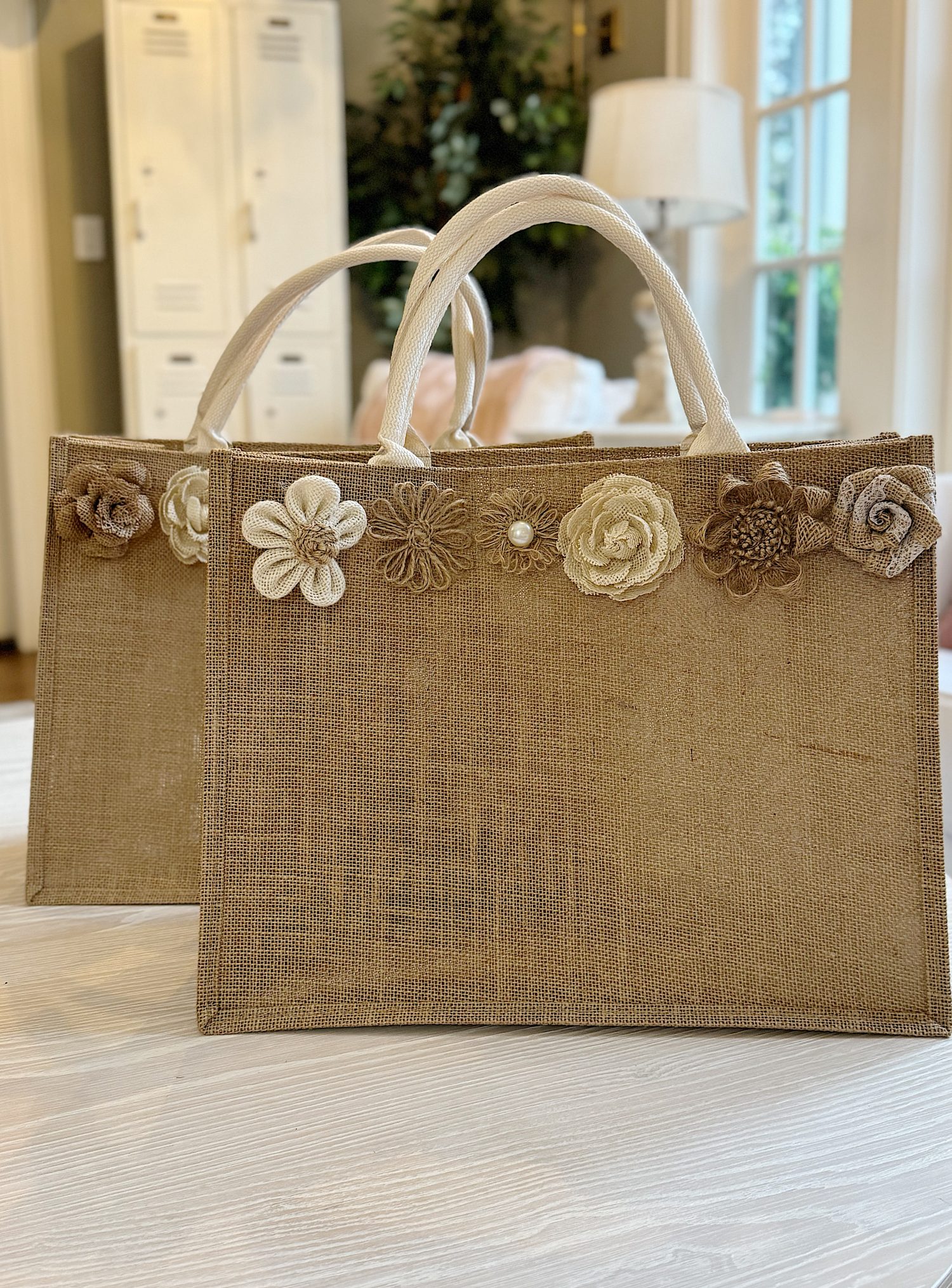 Two brown burlap tote bags with white handles, decorated with fabric flowers in neutral tones, are placed on a light-colored table indoors.
