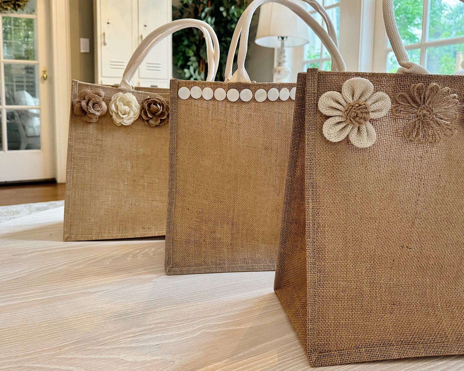 Three burlap tote bags are displayed on a wooden table, each decorated with fabric flowers or circular accents along the top edge.